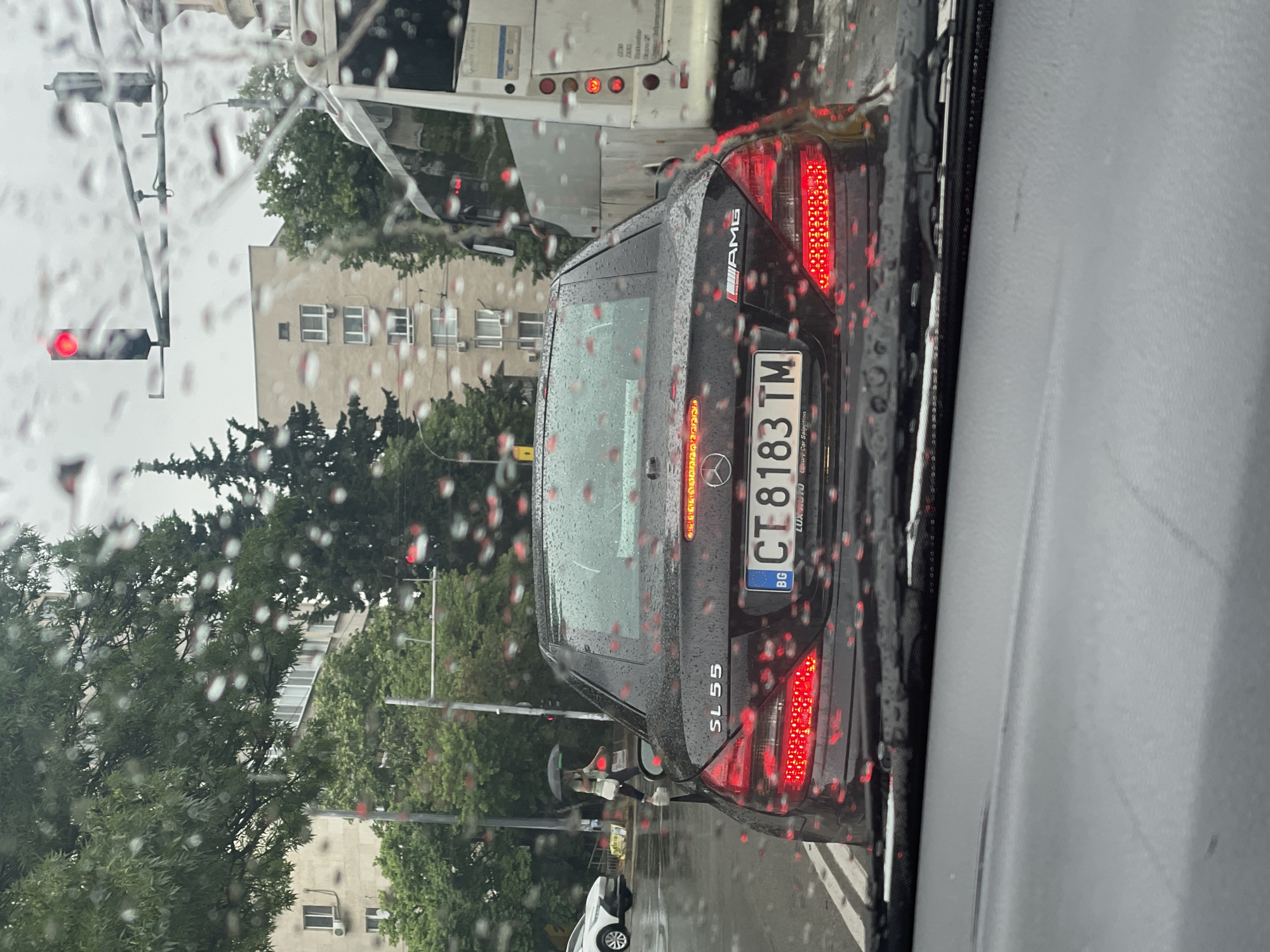 The image captures a scene from inside a vehicle looking forward through a rain-splattered windshield. It is a cloudy, rainy day, evident from the numerous raindrops distorting the view and the overcast lighting.
The primary subject is the rear of a dark-colored Mercedes-Benz SL 55, which is stopped directly ahead. The car's brake lights are illuminated, suggesting it is stopped in traffic, likely at a red light. The vehicle clearly displays an "AMG" badge on the top right of its trunk lid and "SL 55" on the bottom left. Its license plate, positioned in the center, reads "CT 8183 TM" with the country code "BG" (Bulgaria) visible on the left side. Partially visible text on the license plate frame below the numbers appears to say "LUX AUTO".
In front of the Mercedes, a large white vehicle, possibly a bus or truck, is also stopped. Its rear lights are visible. To the far left, a red traffic light is illuminated. On the sidewalk next to the Mercedes, a person holding a black umbrella is visible, walking away from the camera, further emphasizing the rainy weather.
The background shows generic urban or suburban architecture with multi-story buildings and green trees. The wet street surface reflects some light. The overall scene depicts a moment of halted traffic during a rainy day in what appears to be Bulgaria.