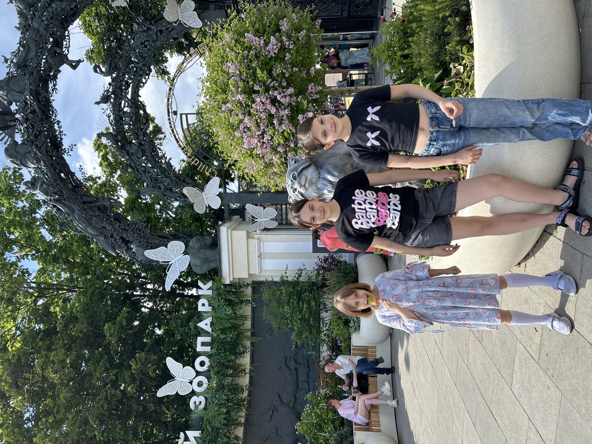 Three girls are captured outdoors, likely at the entrance of a park or zoo, on a bright and sunny day. Two older girls stand in the foreground, smiling towards the camera. One wears a black t-shirt with two white 'X's and blue jeans, while the other wears a black t-shirt with "Barbie Barbie Barbie" text, grey shorts, and sandals. Between them and slightly behind them is a grey statue of a cat.
Further back and to the right, a younger girl in a light-colored dress with pink flamingo patterns and white socks and sandals holds something to her mouth, also looking towards the camera. The background features a large, elaborate metal sculpture forming an archway, intricately designed with tree branches, animals, and several decorative white butterflies. Through the arch, a blue sky with some clouds is visible. Lush green trees and bushes frame the scene, including a flowering bush with light purple blossoms behind the two older girls.
To the left of the scene, white, vertically oriented letters spell out "ЗООПАРК" (ZOO PARK), further indicating the location as a zoo entrance. Another piece of text, "children," is partially visible on a circular element within the metal sculpture. In the far background, two adults are seated on a bench. The overall atmosphere is relaxed and cheerful, suggesting a leisurely visit.