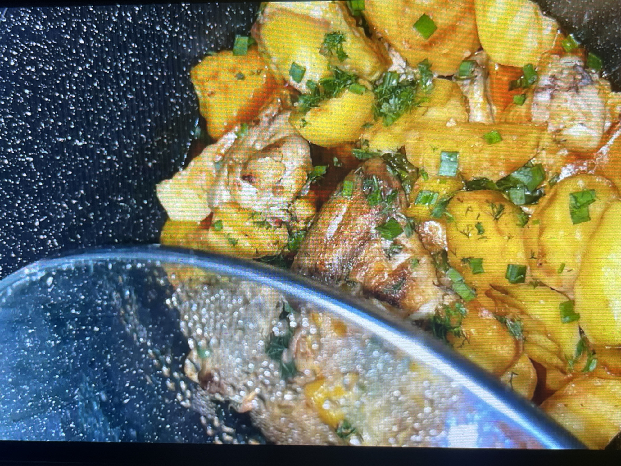 The image shows a close-up, overhead view of a dish being cooked in a pan. The dish consists of pieces of chicken, which appear browned and seasoned, nestled amongst golden-yellow potato wedges. Freshly chopped green herbs, likely dill and green onions, are sprinkled generously over the chicken and potatoes, adding a vibrant touch of green. The food is in a dark-colored pan, and a glass lid, slightly ajar, shows condensation and steam, indicating the dish is actively cooking or has just been finished. The lighting is somewhat harsh, with reflections visible on the glass lid. The scene appears to be a home kitchen setting, with no specific identifiable landmarks of Bishkek City, Kyrgyzstan present.