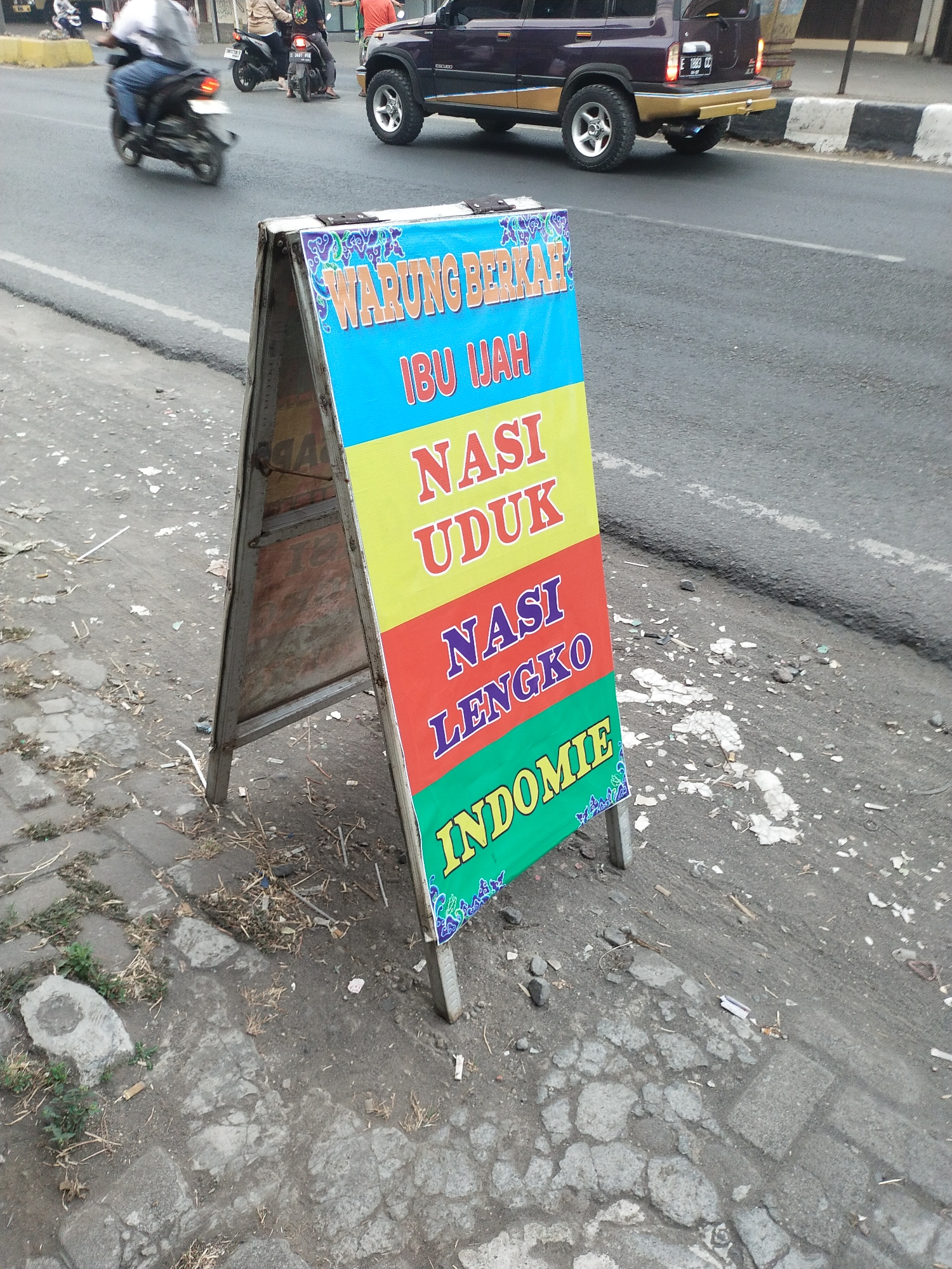 An eye-level, medium-shot photograph captures a street scene in Kemlakagede, Indonesia, dominated by a vibrant A-frame sign advertising food. The sign, positioned on the left foreground, features colorful text on a white background, with blue text reading "WARUNG BERKAH" at the top, followed by "IBU IJAH" in orange. Below this, "NASI UDUK" is written in yellow, and "NASI LENGKO" in red. The bottom section displays "INDOMIE" in green text, adorned with blue decorative swirls. The sign stands on a cracked, gray concrete sidewalk littered with debris, including dry leaves, twigs, and small stones. To the right of the sign, a dark gray asphalt road is visible, with faded white lane markings. A dark purple SUV is parked on the road. Further back, a dark-colored motorcycle carries two passengers, one appearing to be wearing a light-colored shirt. The general environment suggests a typical urban setting with everyday activity.