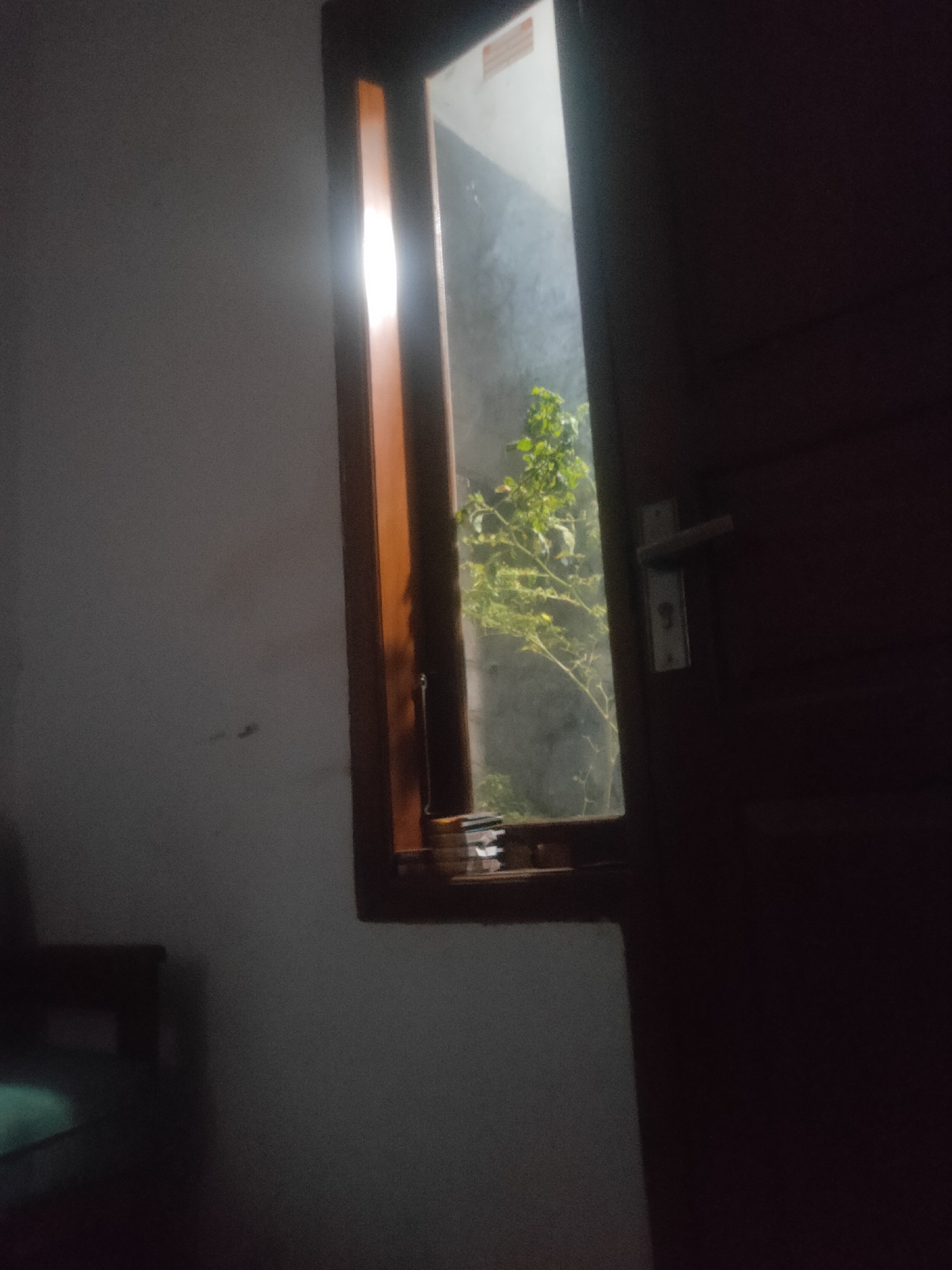 An interior view shows a doorway on the right and a window on the left. The doorway is dark brown and features a silver door handle and lock. The window is framed by a wooden trim. Through the window, a leafy green plant is visible, with light filtering through. The walls are a muted gray. In the foreground, a dark-colored piece of furniture, possibly a chair or sofa, is partially visible on the lower left, with a bright turquoise cushion. A stack of papers is on the windowsill. The scene is dimly lit, suggesting it is nighttime or dusk. The location is Antirogo, Indonesia.