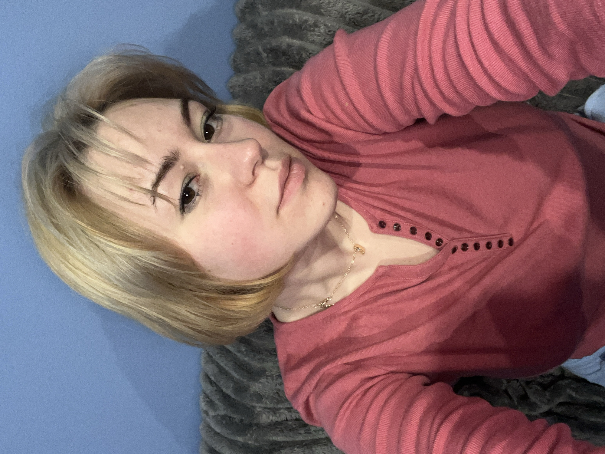 A young woman is captured in a close-up selfie, lying down in an indoor setting in Odesa, Ukraine. Her head is tilted, and she looks directly towards the camera with a neutral to slightly serious expression. She has light blonde hair, parted to the side with bangs across her forehead, and dark eyes with well-defined brows.
She is wearing a long-sleeved, ribbed top in a dusty rose or coral pink color. The top features a Henley-style neckline with a placket adorned with several dark, circular buttons. A delicate gold-colored chain necklace with small charms is visible around her neck.
The background shows two primary elements: a solid light blue surface to her right (viewer's left), which could be a wall or furniture, and a dark grey, plush, textured blanket or throw beneath and to her left (viewer's right). The lighting appears to be soft indoor illumination. No text is visible in the image.