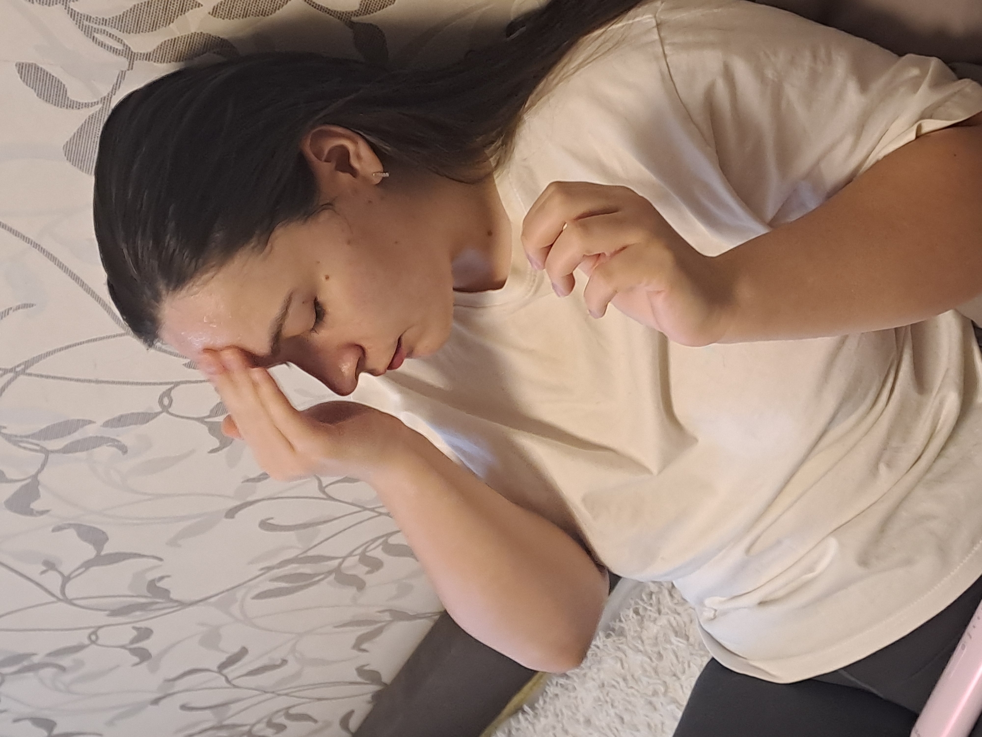 A person is lying on a bed. The person has dark hair and is wearing a white t-shirt and dark leggings. The person's left hand is on their forehead. The bed has a patterned bedspread. The location is unknown.