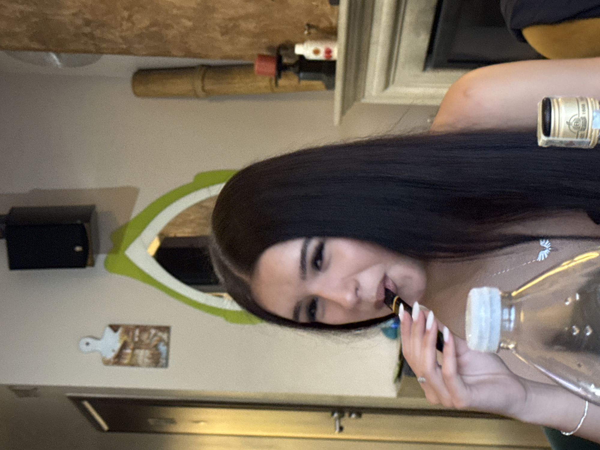 A woman with long, dark hair is holding a water bottle and a small black object, possibly a vape pen, near her mouth. She is wearing a silver necklace and bracelet, and a ring on her finger. The background shows a kitchen setting with a wooden counter, a green-framed mirror, and a small cutting board decoration on the wall. There is also a black speaker on the wall and decorative items above a fireplace. It is not possible to determine from the image if this scene is located in Plovdiv, Bulgaria.