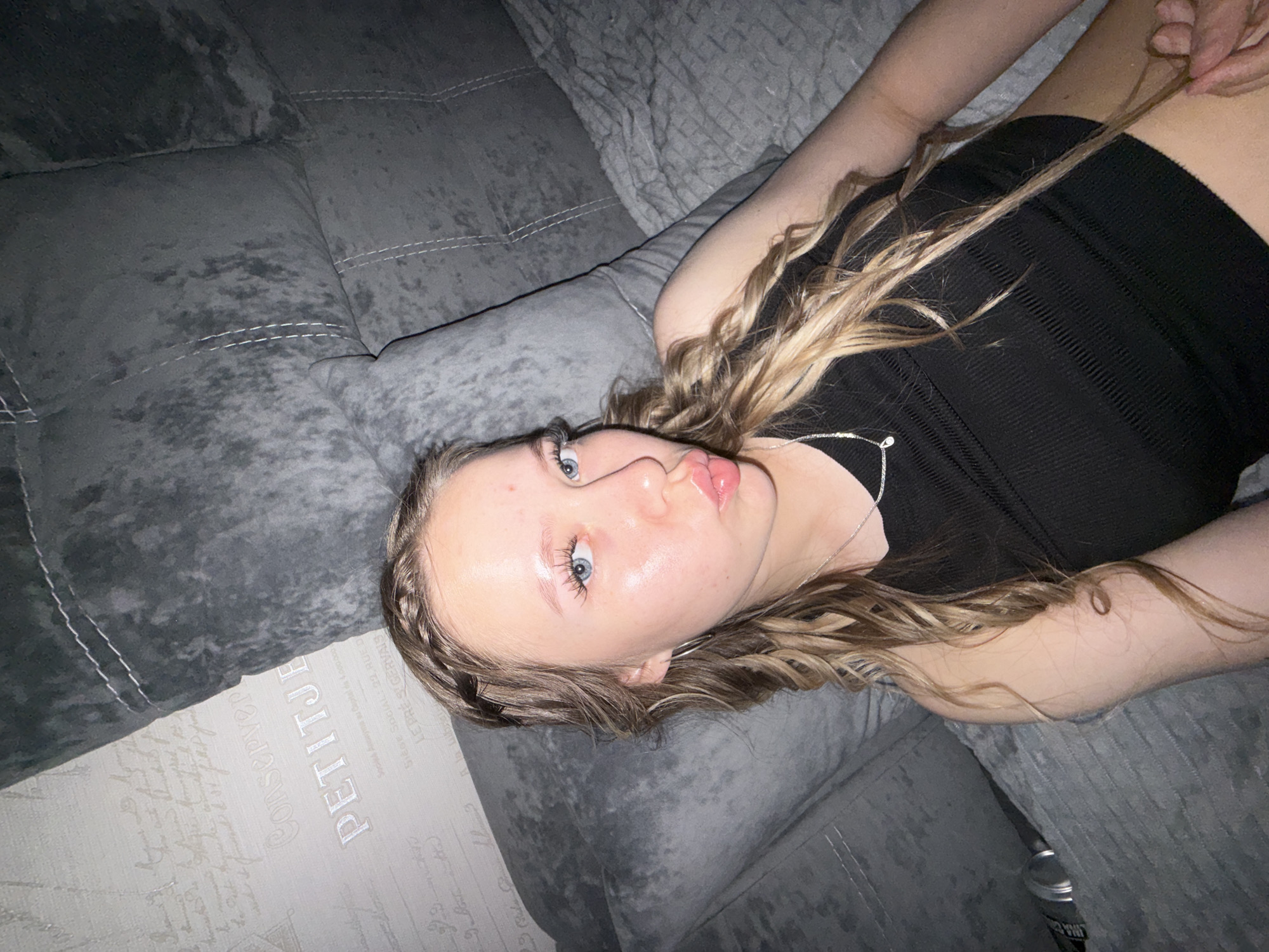 A young woman with blonde hair styled in braids and loose waves is lying down on a gray sofa. She is wearing a black ribbed tank top and a silver necklace with a small pendant. Her eyes are blue, and she is looking towards the camera with her lips pursed in a pout. The background features a gray sofa with stitched detailing and wallpaper with a subtle script pattern. There is no indication of the city of Chaikovsky, Russia.