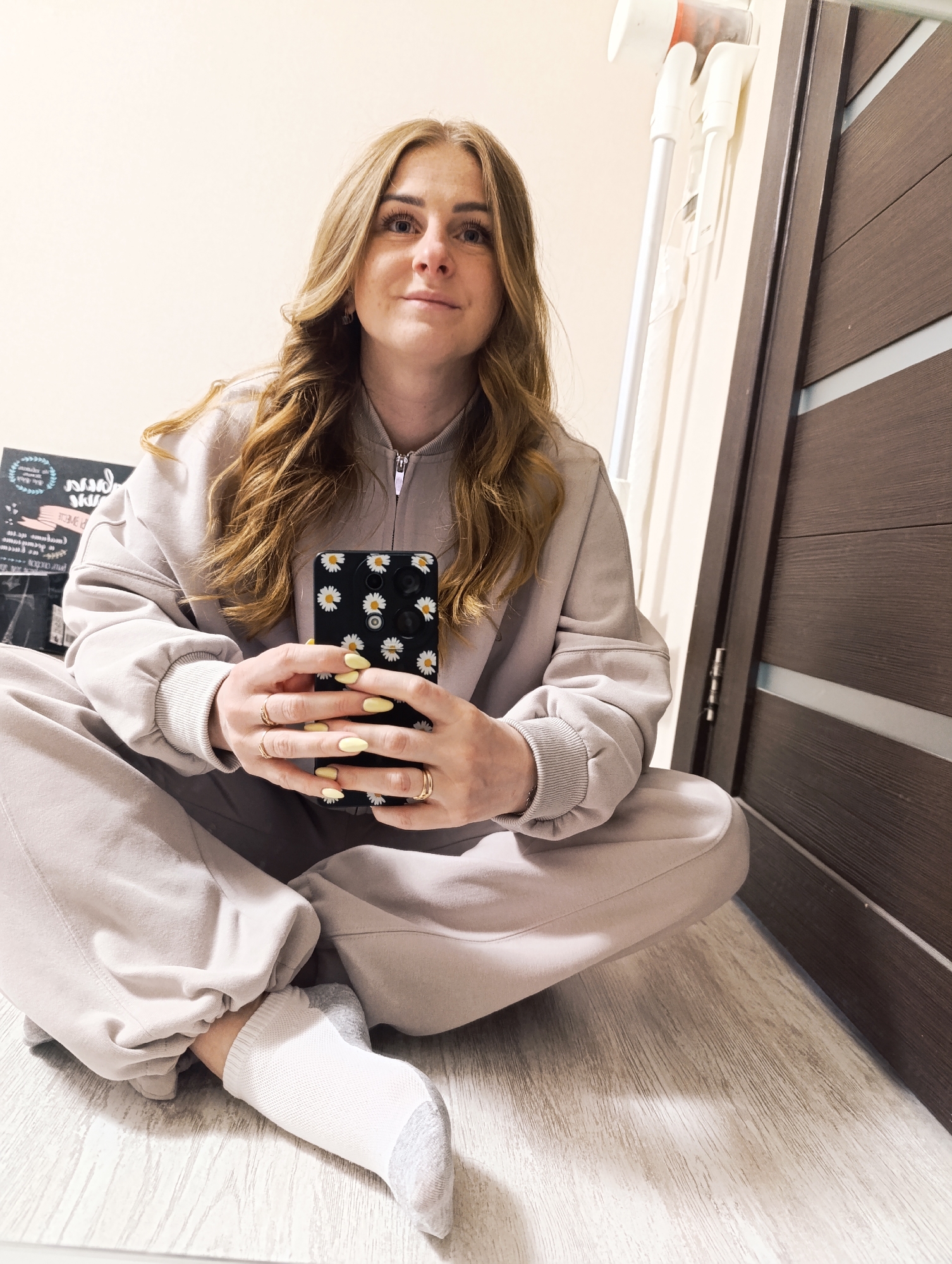 A fair-skinned woman with long, wavy brown hair is seated cross-legged on a light-colored wood-look floor. She is wearing a matching beige sweatshirt and sweatpants, and white socks with gray accents on the toes and heels. The woman is holding a black smartphone adorned with a daisy pattern. Her fingernails are painted yellow. She is looking directly at the camera with a slight smile.
To the right of the woman is a dark brown door with linear detailing. Behind her, against a beige wall, is a white, cordless vacuum cleaner mounted on the wall. To the left is a black box and some plastic bags. The location is an interior space, but the city and region are not discernible.