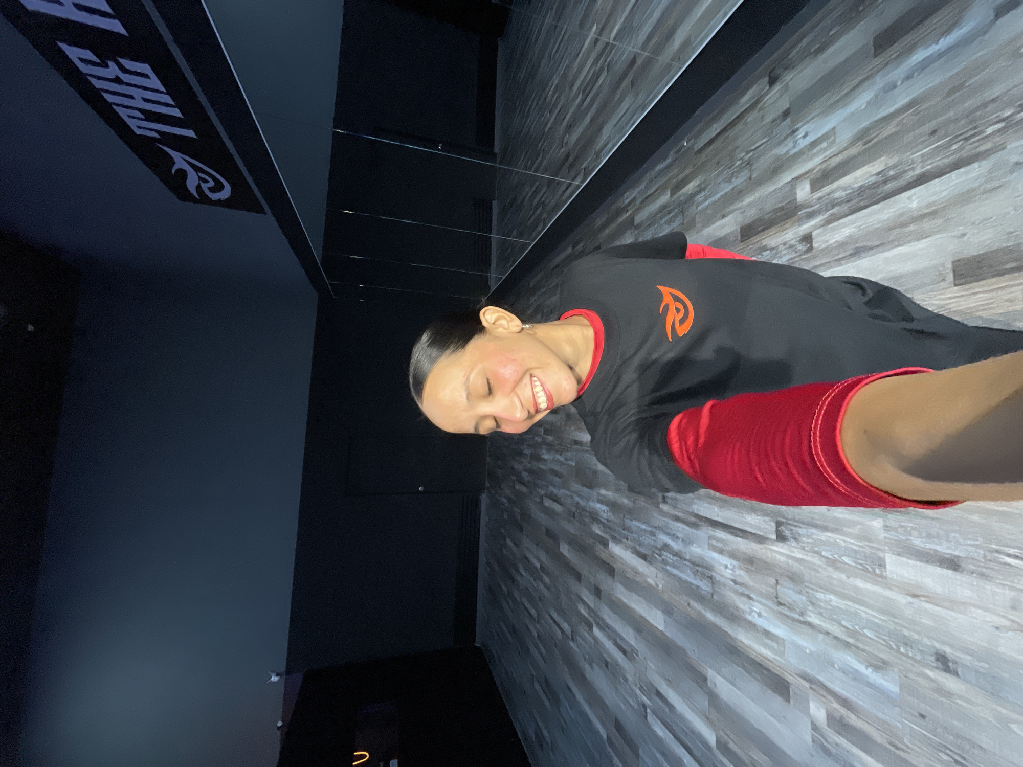 A woman with her eyes closed and a smile on her face is taking a selfie in a dance studio. She is wearing a black t-shirt with an orange logo and a red long-sleeved shirt underneath. The studio has a wooden floor and mirrors on one wall. There is a sign on the wall that reads "THE H". The scene appears to be in a dance studio located in Pampatar, Venezuela.