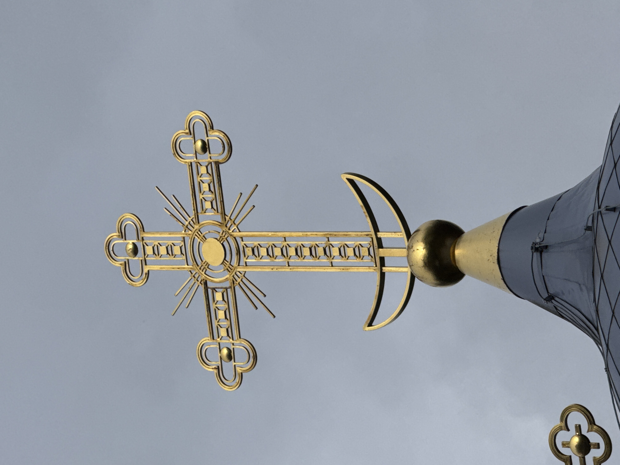 This is a close-up, low-angle shot of a golden Orthodox Christian cross atop a building, possibly a church or chapel, in Neniymaky, Russia. The cross is ornate, with a central circular element surrounded by radiating lines and decorative flourishes at its four ends. A small golden sphere is visible just to the right of the cross, attached to a golden conical structure that is part of the building's roof. This conical structure is dark gray and appears to be made of metal panels. The sky behind the cross is overcast and a uniform light gray, suggesting a cloudy day. The lighting is even, indicating it is likely daytime, though the cloud cover diffuses the sunlight. The image is well-focused on the cross and the adjacent architectural elements, with the background sky out of focus. The overall mood is solemn and serene, emphasizing the religious iconography and the grand scale of the structure.