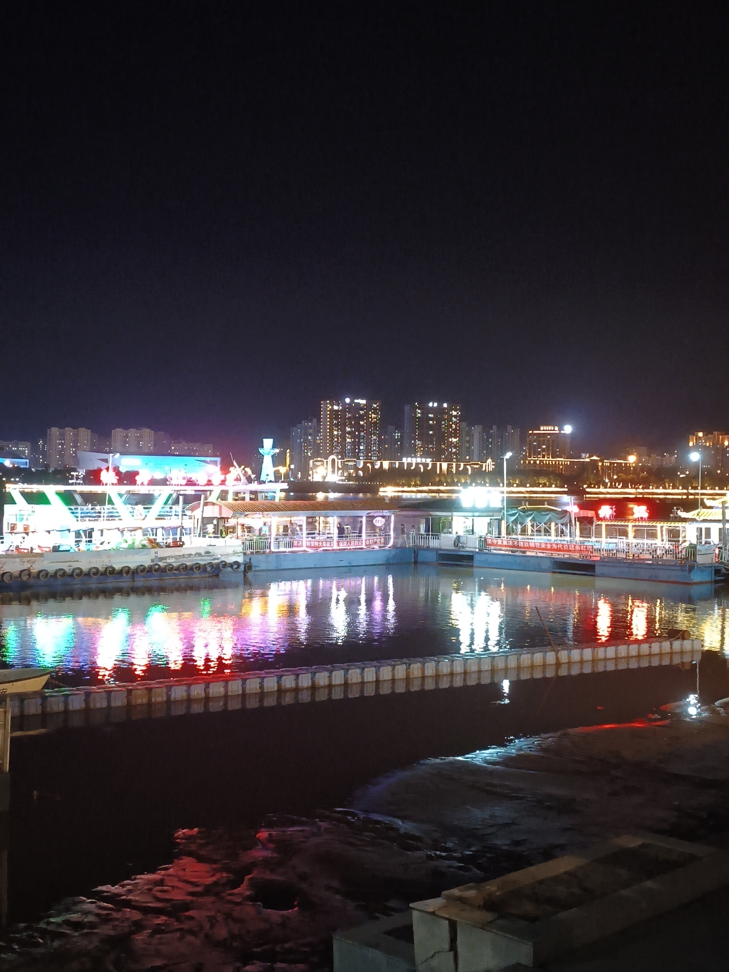 This is a nighttime, long-exposure, vertical-orientation photograph taken from a waterfront. In the foreground, the dark water reflects vibrant colored lights from the structures and buildings in the background. A series of white, rectangular floating structures, resembling buoys or pontoons, are lined up along the edge of the water, creating a border. Beyond these, multiple docked boats or piers are illuminated by colorful lights in green, pink, and red, casting bright reflections on the water's surface. The boats appear to be multi-tiered and are adorned with various lights. In the background, the cityscape is visible with numerous tall buildings, many of which are lit up with windows. A prominent white structure, possibly a monument or decorative element, stands out among the buildings. The sky is dark and unlit, indicating nighttime. The image was taken in the city of Dongxing Street, China.