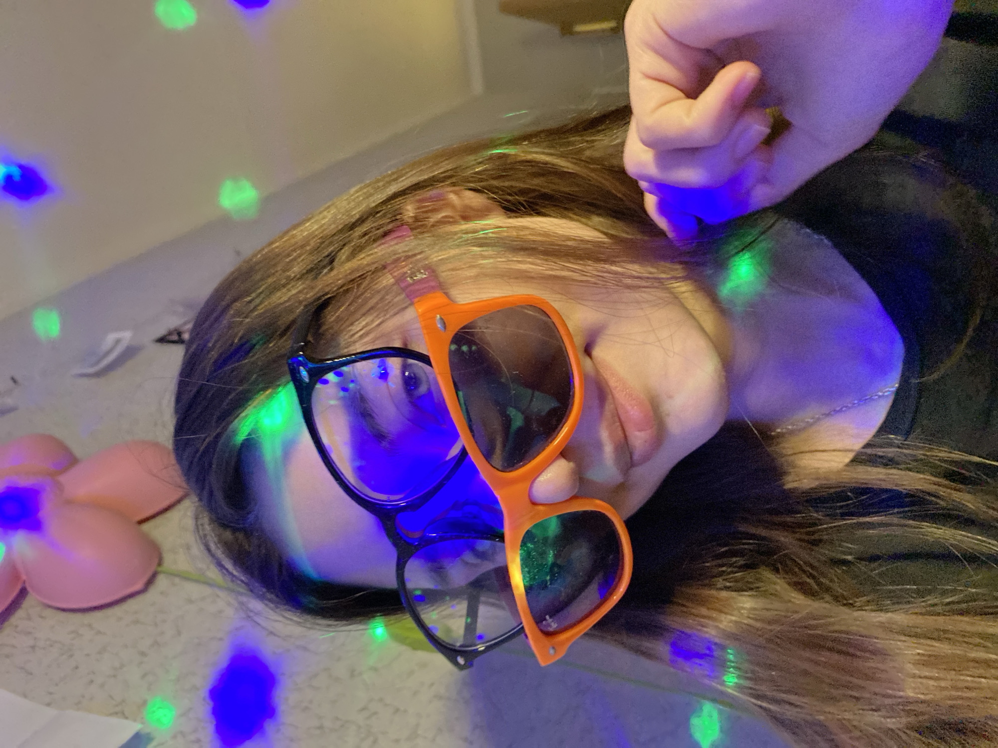 A person with long brown hair is lying down, wearing two pairs of glasses—one black and one orange. Their hand is near their head. The scene is illuminated with blue and green lights. There is a pink flower-shaped object next to the person. The location is unknown and there is no indication this is in Votkinsk, Russia.