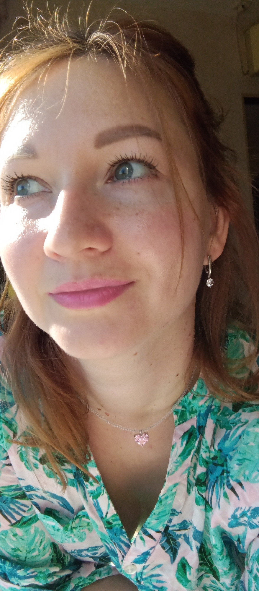 A close-up shot shows a fair-skinned woman with reddish-blonde hair and blue eyes. She has freckles and is wearing pink lipstick. Her expression is a slight smile, and her gaze is directed upwards and to the left. The bright sunlight accentuates the highlights in her hair and casts shadows across her face. She is wearing a silver necklace with a pink heart pendant and matching earrings. She is also wearing a green and pink floral-patterned shirt. The location appears to be indoors near a window. The city is Unknown, Unknown.