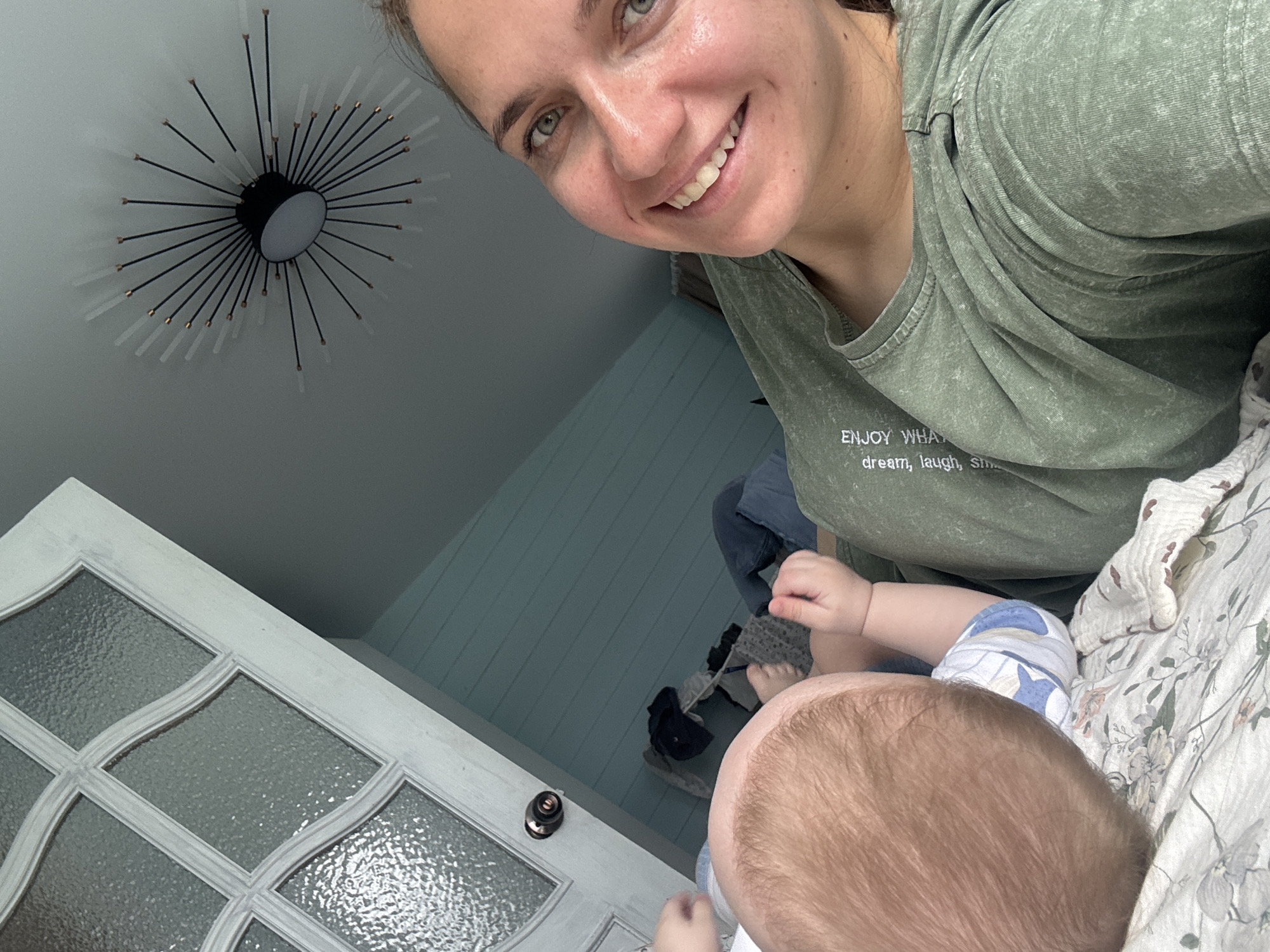 An intimate indoor scene in Раздолье, Russia, captures a smiling woman and an infant from an overhead perspective. The woman, with light skin, green eyes, and light brown hair pulled back, is featured in the upper right quadrant of the image, smiling broadly at the camera. She wears an olive green t-shirt with text visible on the chest that reads "ENJOY WHAT" and "dream, laugh, smile."
Below her, the back of an infant's head with light brown hair is visible. The baby appears to be lying down, possibly on a floral-patterned blanket or sheet, and is seen wearing a blue and white patterned onesie on its arm. The baby seems to be holding a bottle.
The setting includes a contemporary sputnik-style ceiling light fixture in the upper left, featuring a black central disc with numerous radiating black rods ending in frosted tubular bulbs. To the left, a white painted door with multiple textured glass panes and a dark doorknob adds architectural detail. A light blue or grey planked wall is visible in the background, creating a cozy atmosphere. The lighting suggests it is daytime. The woman's joyful expression conveys a happy and tender moment.