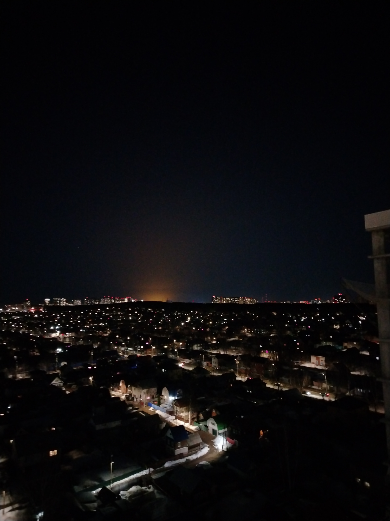 The image captures a nighttime view of the city of Izhevsk, Russia, from an elevated perspective. In the foreground, rooftops of houses covered in snow are visible. Streetlights illuminate the roads and pathways, adding to the overall glow of the city. In the distance, the city skyline features clusters of lit-up buildings and structures, creating a sense of urban development. The night sky above is dark. A diffuse glow on the horizon suggests the presence of city lights further in the distance. Construction is visible on the right side of the image.