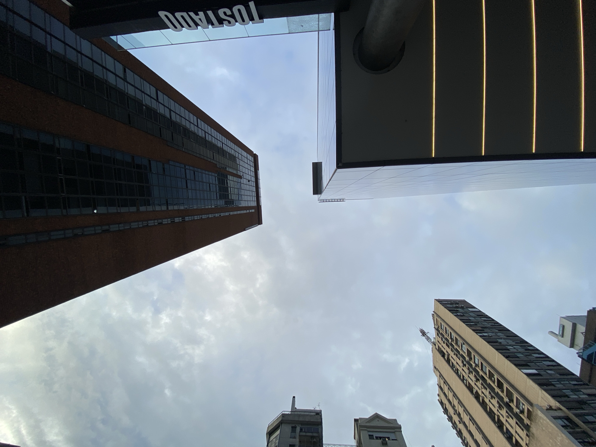 This image depicts a dense urban skyscape, viewed directly upwards from street level under an overcast sky. The central area is dominated by a mostly grey, cloudy sky. Surrounding the sky are portions of multiple tall buildings.
On the upper side, extending diagonally from top-left to top-right, is a modern high-rise building characterized by a brown brick facade interspersed with dark, rectangular glass windows forming a grid pattern.
To the right, a contemporary building with a dark, reflective glass exterior is visible. A vertical sign bearing the white capital letters "TOSTADO" is prominently displayed on its side, reflecting portions of the sky. Below this sign, the building features multiple horizontal illuminated yellow strips. A large, grey cylindrical column or pipe is partially visible at the bottom-right corner, extending upwards alongside this building.
On the lower-left, a tall, light-colored (possibly beige or concrete) residential tower rises. This building exhibits numerous rectangular windows across its facade, and multiple external air conditioning units are visible protruding from some windows. A portion of a construction crane's jib is visible at the very top of this residential tower. Further to the far-left, partial views of smaller, older, light-colored buildings are discernible.
No people, discernible actions, or interactions are present in the scene. The architecture and the specific "TOSTADO" signage indicate the location is in the vicinity of **Diagonal Roque Sáenz Peña (Diagonal Norte), 943, within the San Nicolás neighborhood of Buenos Aires, Argentina.**