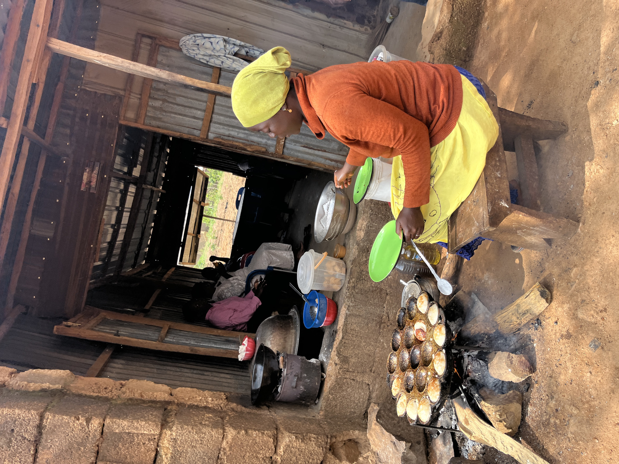 In Barakin Akawo, Nigeria, a woman is cooking small cakes in a traditional outdoor kitchen. The scene is rustic, with a stone wall on the left and corrugated metal walls and a wooden roof forming the structure of the kitchen. The woman, wearing a yellow headscarf and an orange sweater over a yellow skirt, is seated on a wooden stool, actively tending to a circular pan filled with cooking cakes over an open fire. The pan has many indentations, each holding a small cake, some of which are golden brown, indicating they are nearing completion. In the background, other people are visible sitting inside the structure, suggesting a communal or family setting. Various cooking utensils and containers, including bowls, a sieve, and a larger pot, are scattered around the cooking area, indicating a functional food preparation space. The ground is composed of reddish earth and stones.
