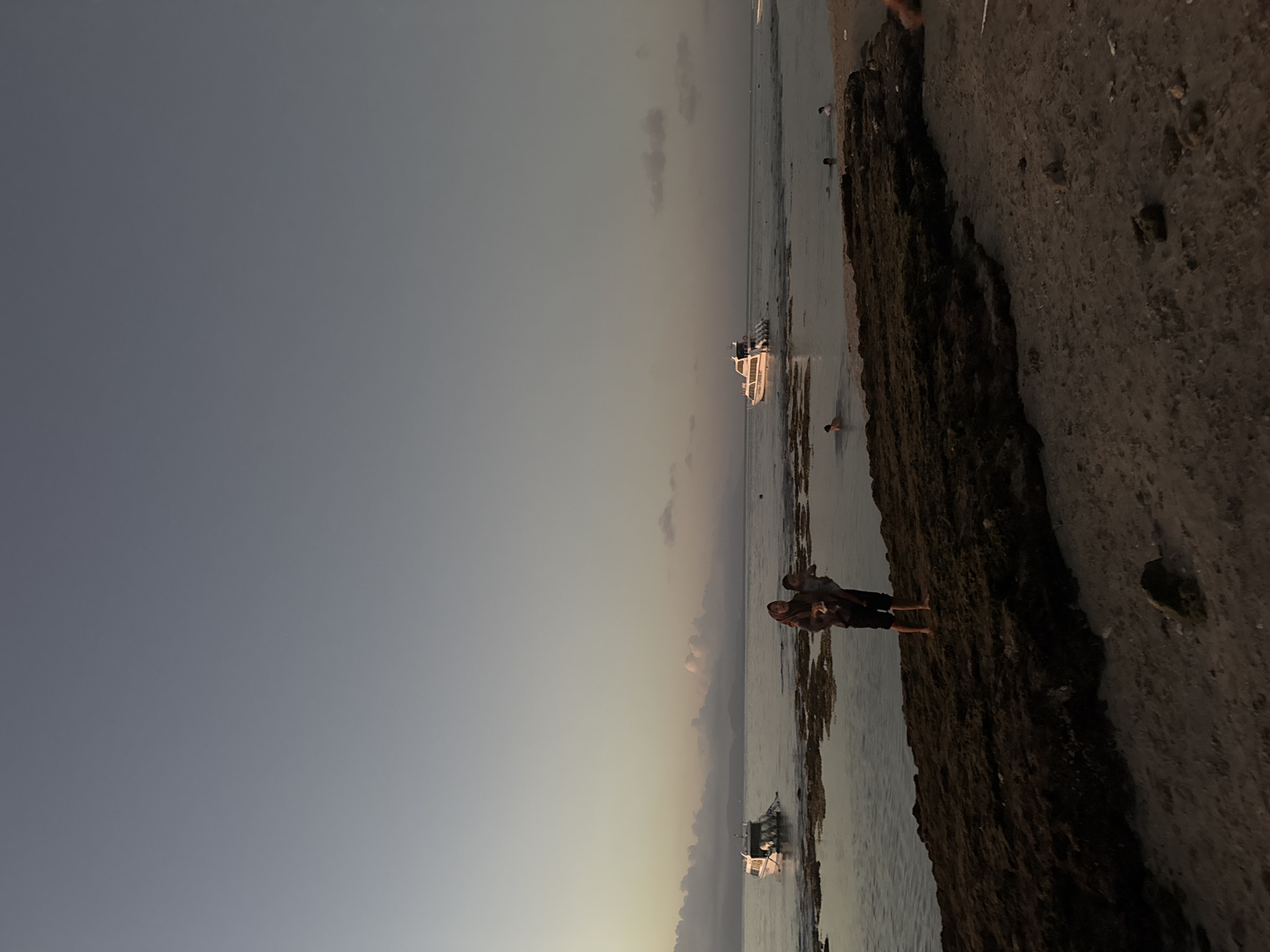 The image depicts a serene beach scene in Nusa Penida, Indonesia, likely at dusk. The sky is a gradient of soft blues and grays, transitioning into a pale yellow near the horizon, suggesting the setting sun. The calm ocean gently laps against a rocky shoreline.
In the foreground, two people are standing on the dark, textured rocks near the water's edge. They appear to be a couple, with one person embracing the other from behind. Their poses suggest a relaxed and intimate moment. Further out in the water, a white boat with a few passengers is visible, and another smaller boat is anchored closer to the shore. A few other individuals are wading in the shallow water.
The overall atmosphere is peaceful and tranquil. The lighting is soft, casting a warm glow on the scene. The location is identifiable as Nusa Penida by the distinctive rocky formations and the type of boats commonly seen in the region. The water appears clear and shallow in many areas. There is no visible text in the image.