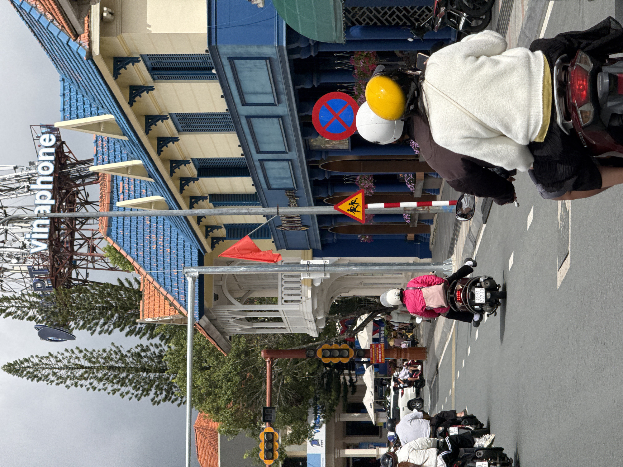 This image depicts a street scene in Da Lat, Vietnam, likely a city center or main thoroughfare. The foreground features several motorcycles with riders wearing helmets, navigating a paved road with white lane markings. One motorcycle in the immediate foreground carries two individuals; the rear passenger wears a white fuzzy sweater and a yellow helmet. Behind them, other motorcycles and riders are visible, including a rider in a pink jacket.
The background reveals distinct architecture. A building with a bright blue tiled roof and pale yellow walls, adorned with decorative white trim and dark blue shutters, lines the street. A white balcony with ornate railings extends from a section of this building. A red flag is attached to a pole near the building. Further in the distance, tall pine trees are prominent, and a large metal telecommunications tower with a "Vinaphone" sign is visible against a cloudy sky. Traffic signals, including a multi-light signal and a pedestrian crossing signal with stick figures, are present on a traffic pole. A red and white striped pole with traffic signs, including a "no parking" symbol with a blue cross, is also visible. The overall atmosphere suggests a busy urban environment with a blend of modern infrastructure and traditional Vietnamese architectural influences.