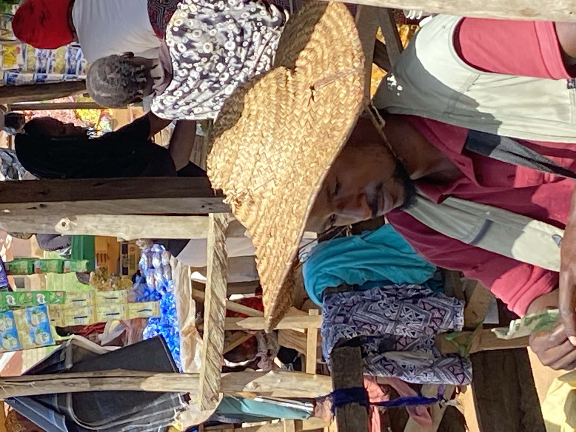 The image depicts an outdoor marketplace in Riyom, Nigeria. A man wearing a straw hat, red shirt, and a light-colored vest stands near a wooden structure used to display items for sale. Stacks of merchandise, including boxed goods and bundled clothing, are visible behind the man. Other people are present in the market, browsing the goods.