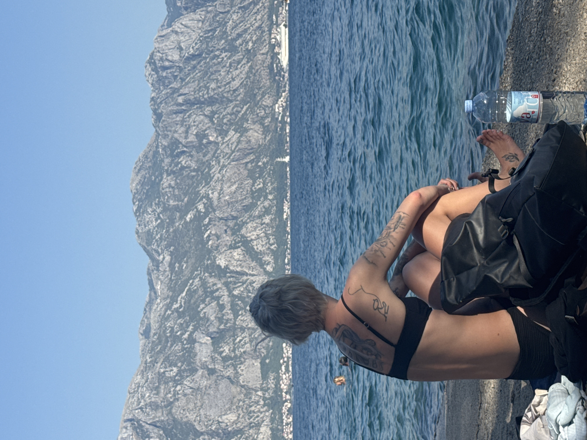 A person with short grey hair and tattoos on their back and arms sits facing away from the camera, looking out at the sea in Perast, Montenegro. They are wearing a black bikini top and bottoms. Next to them is a black backpack and a bottle of water. In the background, the bay of Kotor is visible, with mountains rising in the distance. There are a few other people in the water.