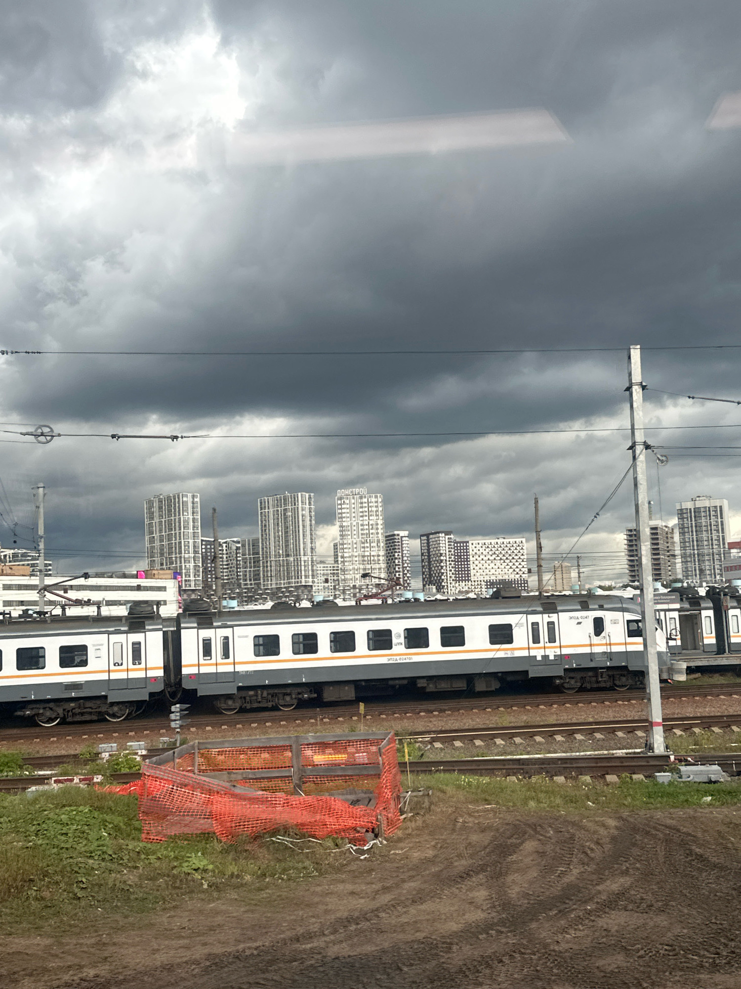 The image shows a train at a railway station in Moscow, Russia, under a cloudy sky. The train is white and gray with an orange stripe, and it is parked on a set of train tracks. In the background, there are several tall buildings, some of which appear to be under construction. There is also an orange construction fence in the foreground, and a patch of green grass and dirt. The overall scene suggests an urban environment with ongoing development.