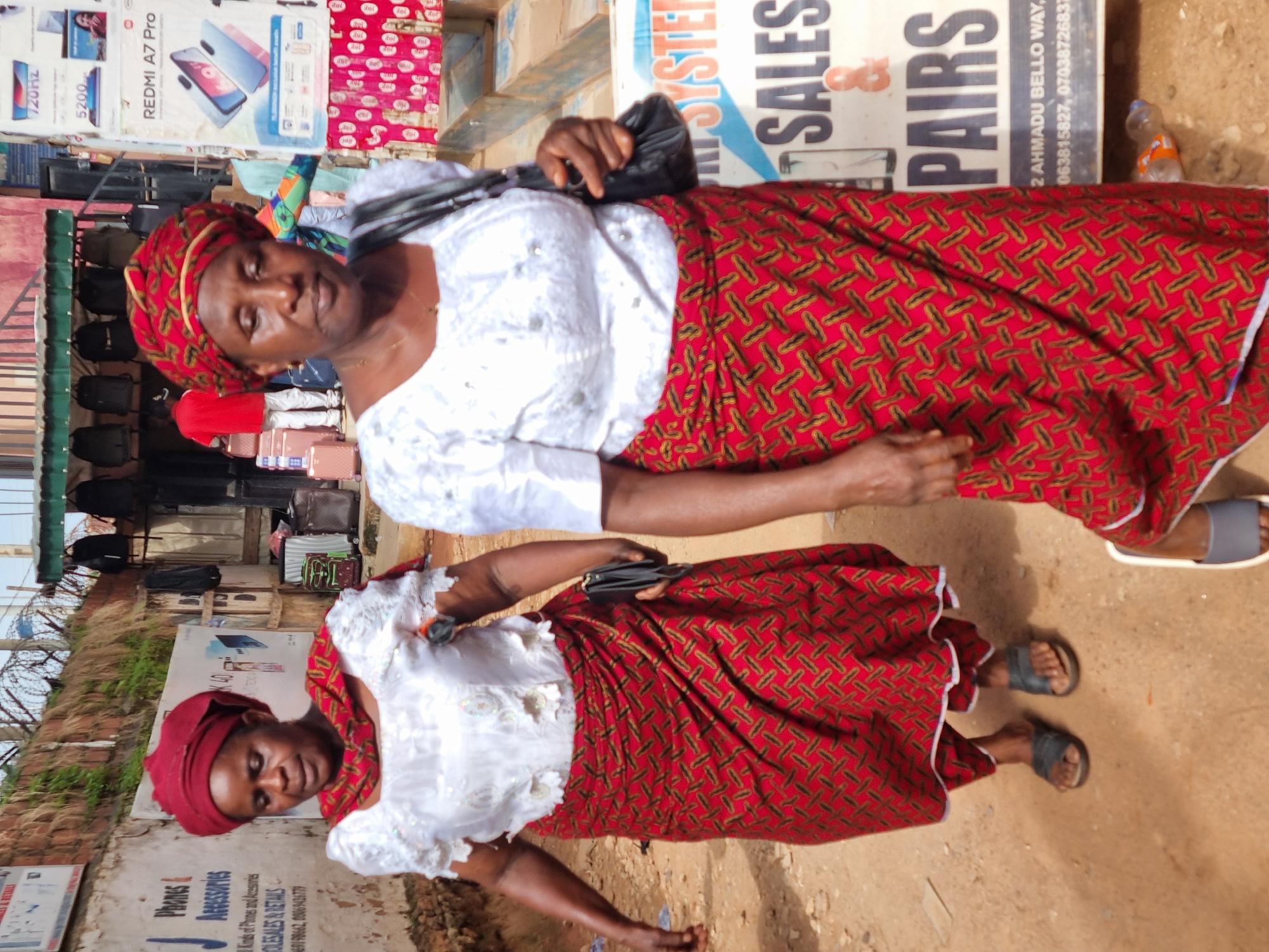 Two women dressed in traditional African attire walk in an outdoor marketplace. The woman in the foreground wears a white, embroidered top and a red and gold patterned wrap skirt. A black handbag is slung over her shoulder. The woman behind her wears a similar outfit, with a red headscarf and a red and gold patterned wrap skirt. They appear to be walking away from the camera. In the background, stalls are visible, displaying various goods, including phones and accessories. The environment suggests a busy urban setting, possibly in Jos, Nigeria, indicated by the signs for "Ahmadu Bello Way." The ground is unpaved, with some scattered debris. The lighting indicates it is daytime.