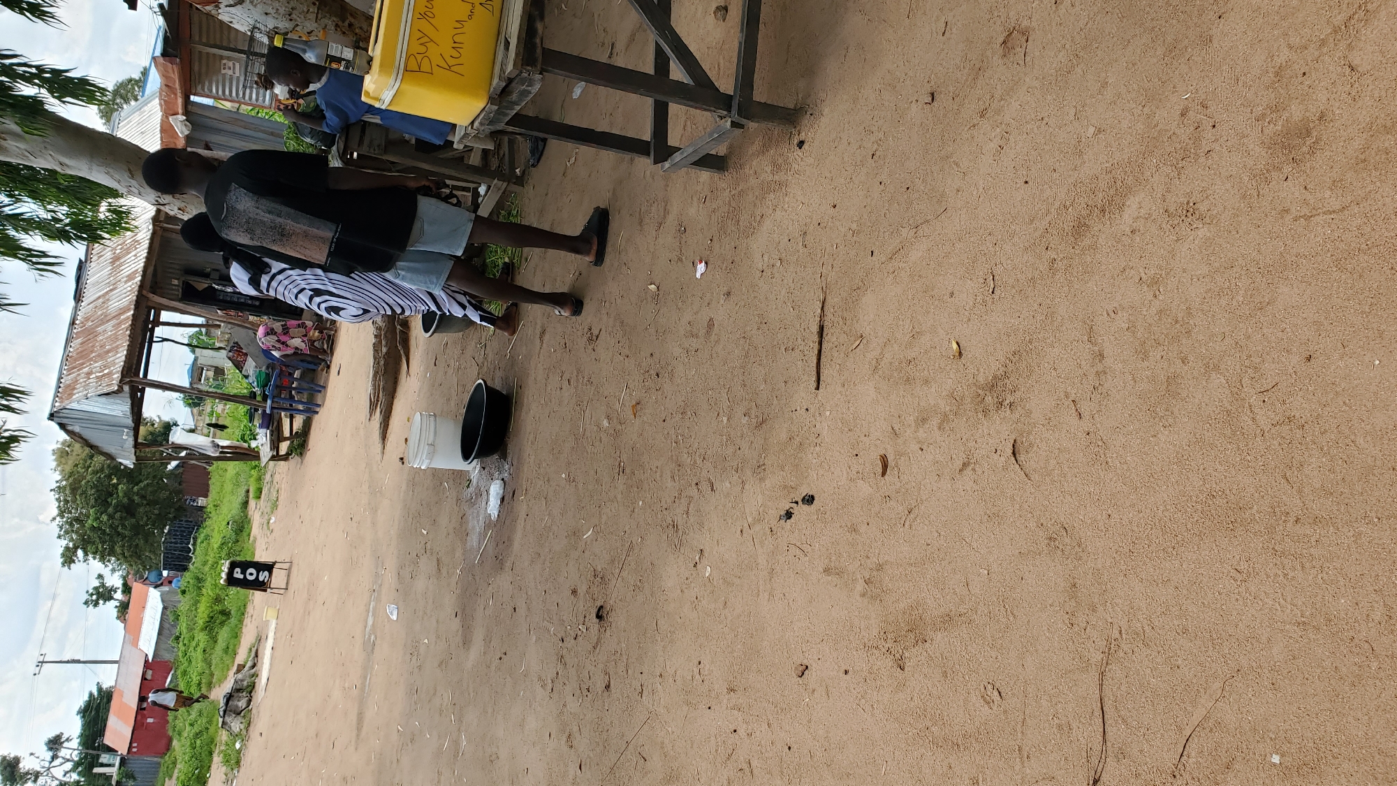 In Jalingo, Nigeria, on a dirt road, a group of individuals are gathered near a makeshift stall. A young boy in a blue shirt is interacting with someone behind a yellow cooler. To the left, a young man in a black t-shirt and light blue shorts stands with his back to the camera. Another person wearing a black and white patterned dress is also present. In the background, there are several simple structures, including a red building and a stall with a corrugated metal roof. A sign displaying "POS" with a logo underneath is visible on the left side of the scene, indicating a point of sale or service. The ground is sandy and dusty, with scattered debris. The overall setting appears to be an informal market or roadside vendor area.