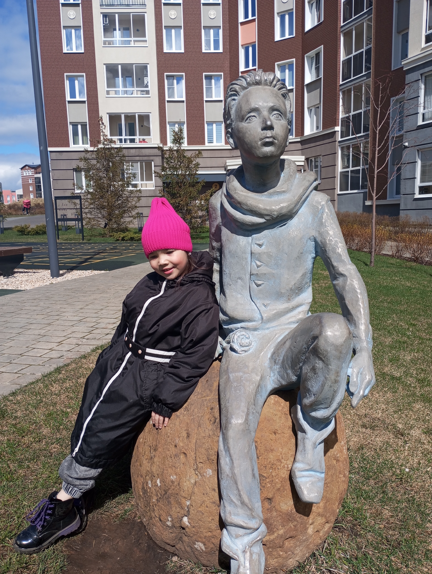 A young girl in a pink beanie and black snowsuit leans against a statue of a boy sitting on a large rock. The statue is weathered and appears to be made of metal. The girl is smiling slightly at the camera. They are positioned on a grassy area with paved walkways nearby. In the background, there are modern apartment buildings with brick and light-colored facades, located in Pervomaysky, Russia.