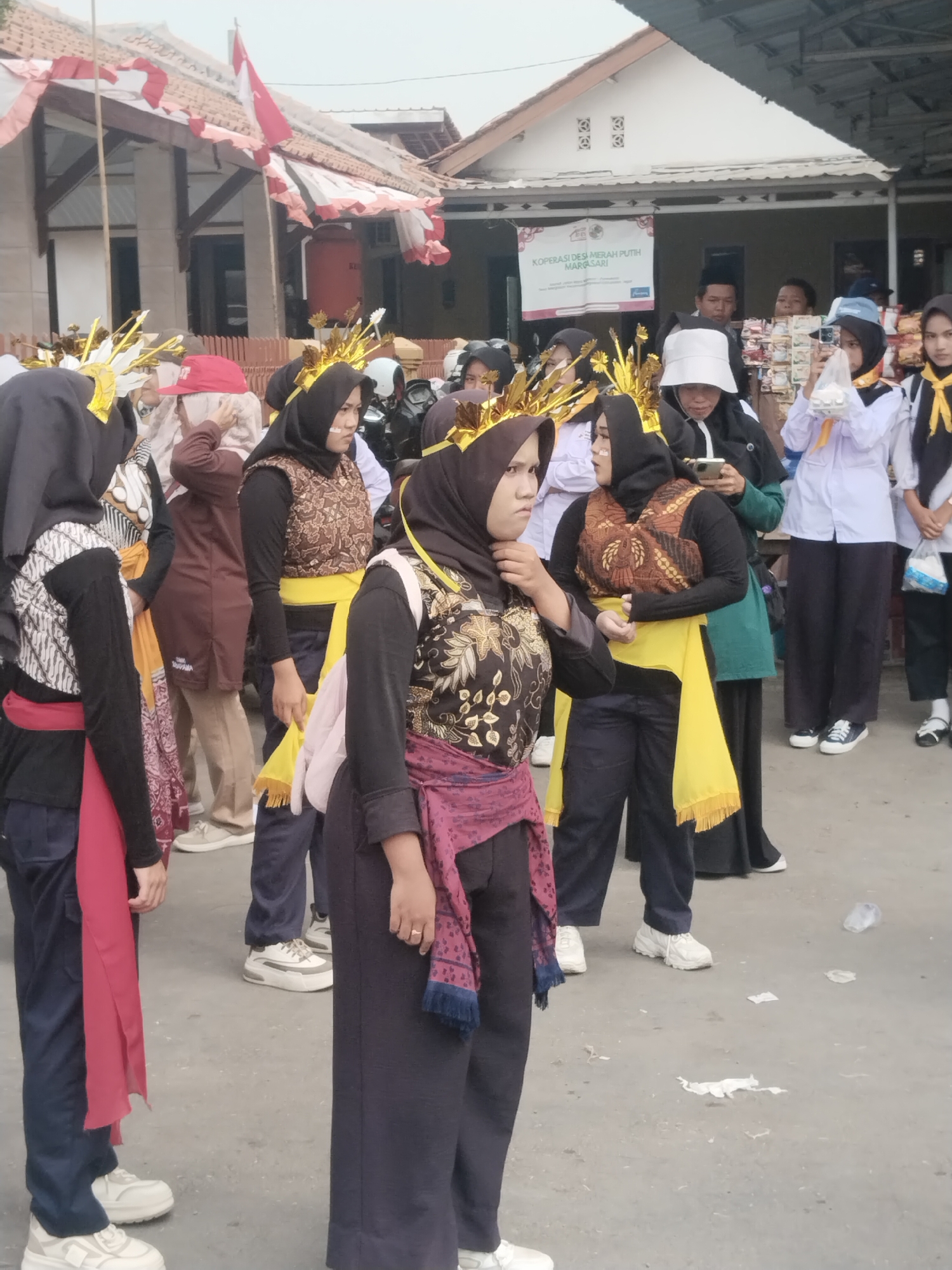 In Margasari, Indonesia, a group of women dressed in traditional attire participate in an outdoor event. The women are wearing hijabs and ornate gold headpieces, with some sporting traditional batik vests over long-sleeved tops and pants. They are adorned with brightly colored sashes, including red and yellow, adding to the festive atmosphere. In the background, a banner that reads "KOPERASI DESA MERAH PUTIH MARGASARI" is visible, indicating the event's affiliation with a local cooperative. The setting appears to be a street or open area, with buildings and a fence in the background. Some individuals are holding cameras, suggesting the event is being documented. The ground is a rough concrete surface with scattered litter, including plastic bottles and paper. The overall scene depicts a cultural gathering or procession within the community of Margasari.