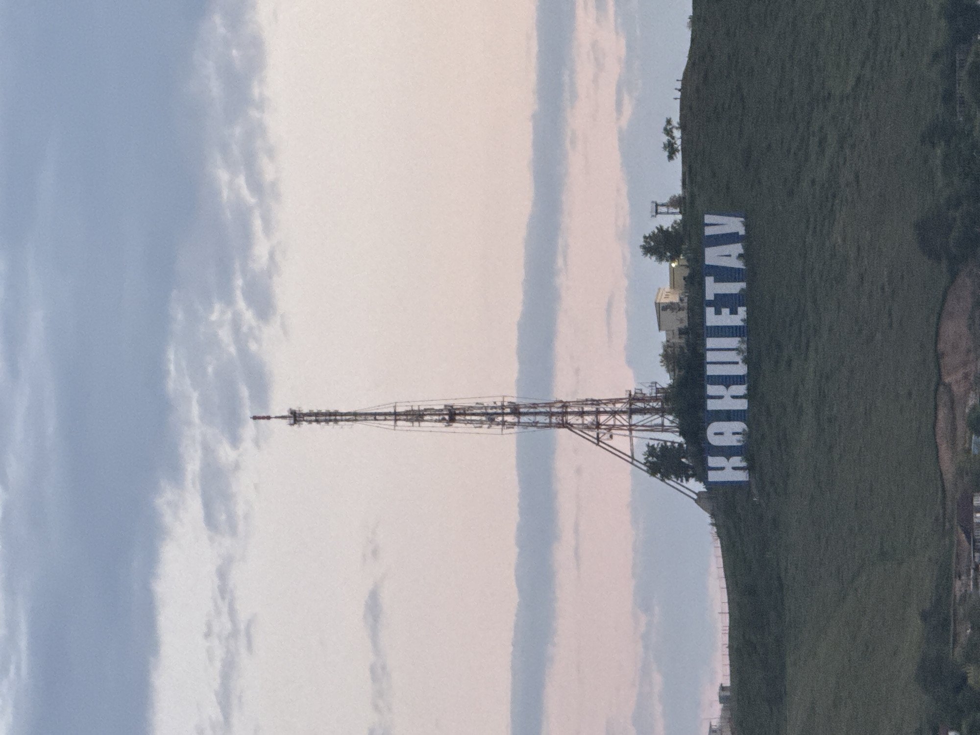 The image shows a telecommunications tower in Kokshetau, Kazakhstan, and the hillside upon which the name of the city is written in large Cyrillic letters. The sky is cloudy with an even light. The hillside with the Kokshetau letters is covered in green grass. Some trees are visible. Buildings can be seen in the distance.