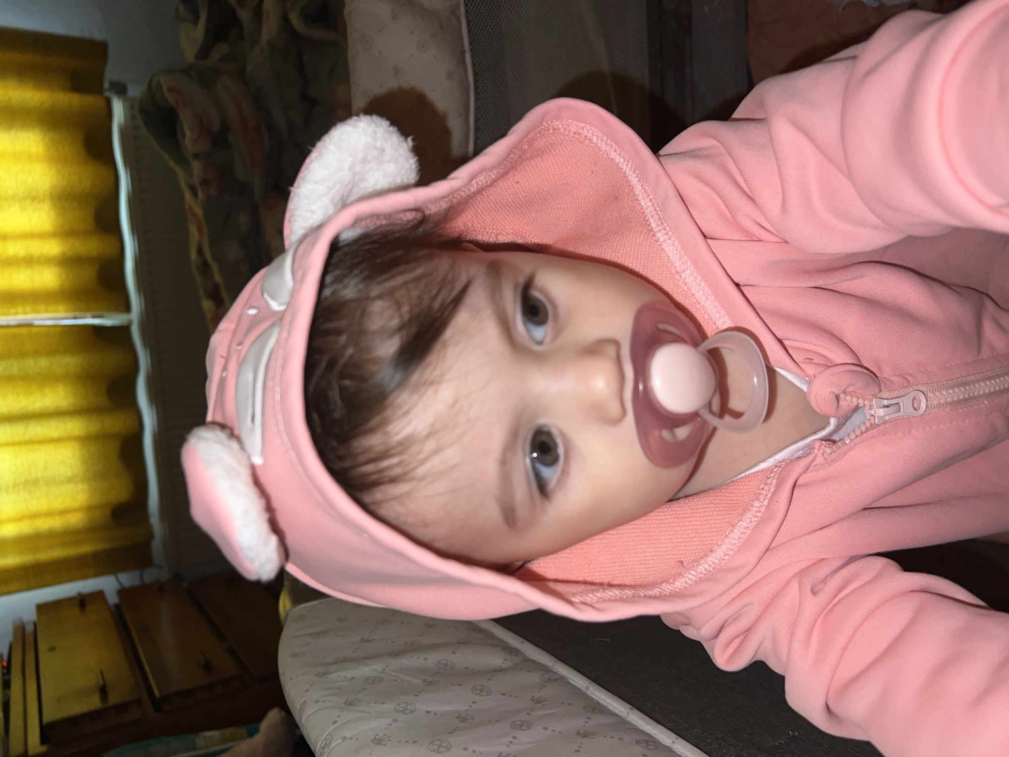 A baby wearing a pink hooded sweatshirt with white furry ears and a pink pacifier in their mouth is lying on their back in what appears to be a crib or playpen with a patterned mattress. The location cannot be determined to be Elena, Bulgaria. There are brown and yellow blinds in the background to the left. The baby is looking at the camera. The photo is taken from a slightly above and angled perspective. A zipper is visible on the sweatshirt.