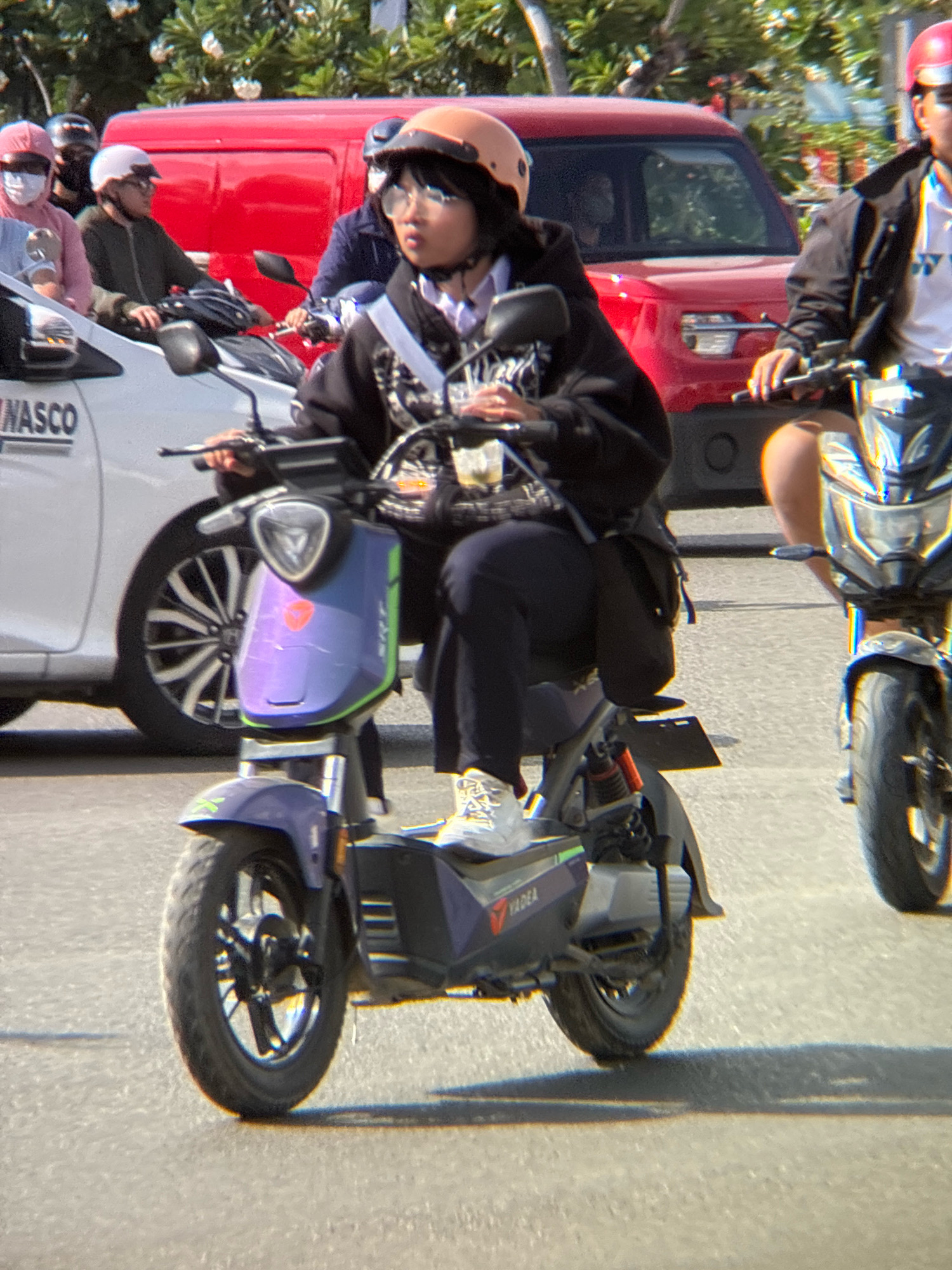 The image depicts a street scene in Nha Trang, Vietnam, with several individuals on motorcycles and a car. In the foreground, a young person wearing a black hoodie, dark pants, white sneakers, and an orange helmet is riding a purple and green electric scooter. They are looking to the right. To their right, another person wearing a black jacket and a red helmet is riding a motorcycle. To the left of the scooter rider, a white car with "NASCO" visible on its side is parked. In the background, a red van and other vehicles are present, along with some trees. The scene appears to be a typical daytime street in a bustling urban environment.