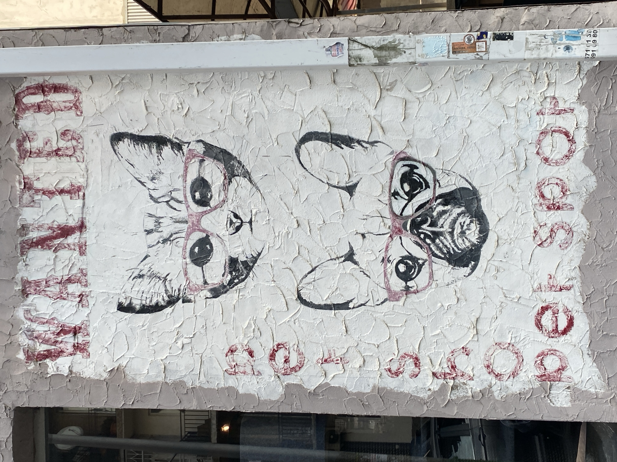 A mural of a cat and a French bulldog wearing glasses is painted on a textured wall. The mural reads "WANTED" at the top, and "PET SHOP" vertically on the left side, painted in red. The mural is located on the exterior wall of a building in Batumi, Georgia. The wall is cracked and weathered. There is a window to the left of the mural, showing the interior of a building and a parked car. A white pole is to the right of the mural with stickers on it, and some fencing is visible in the background.