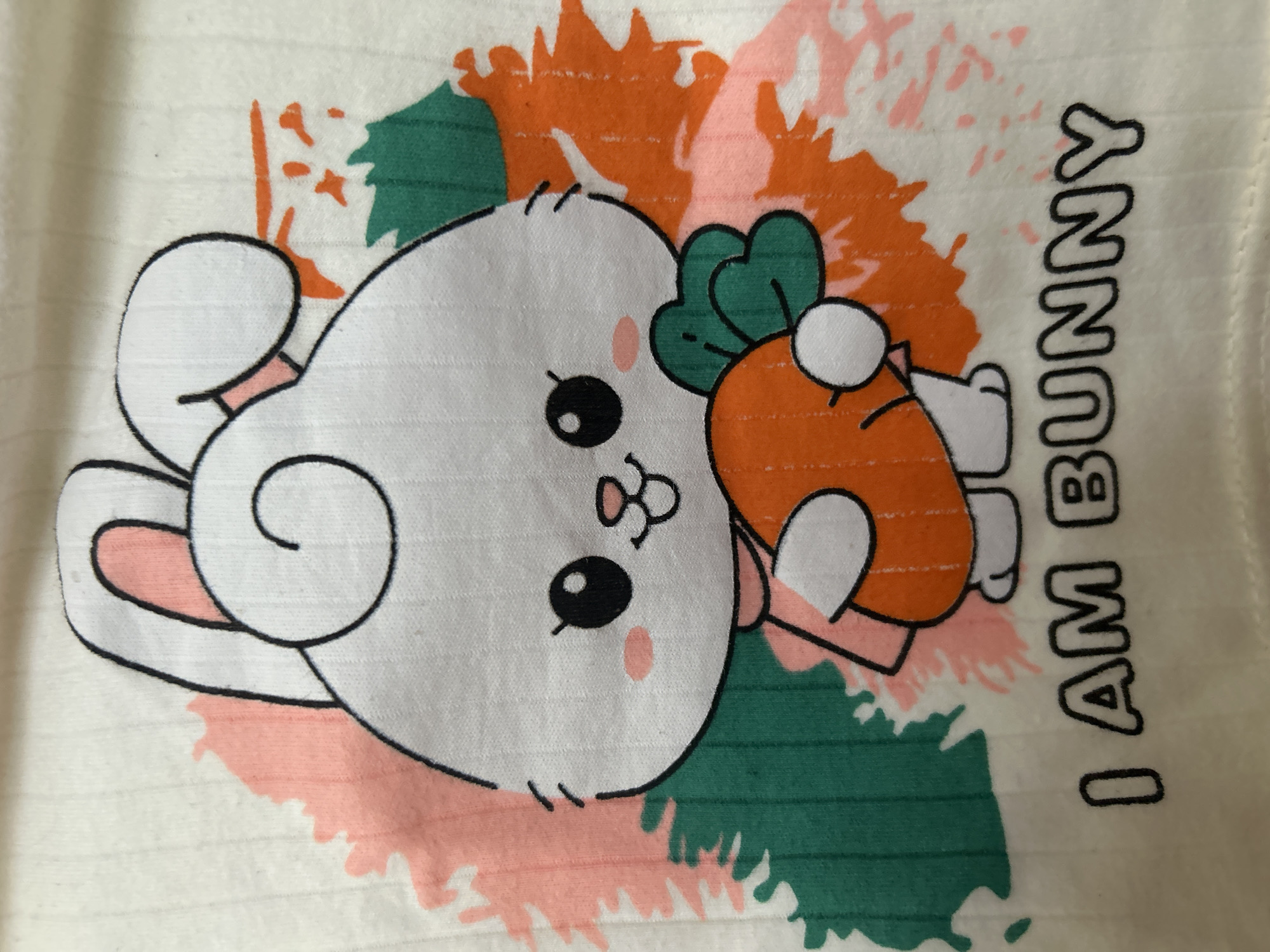 The image presents a close-up, rotated view of a light-colored, possibly off-white, fabric, likely a garment, featuring a printed cartoon design. The central subject is a cute, stylized white bunny character. The bunny has large black eyes with delicate eyelashes, small pink blush marks on its cheeks, a tiny pink nose, and two small top teeth. One of its ears is prominently displayed as a black-outlined white spiral, while another ear is partially visible behind its head. The bunny is depicted holding an orange cartoon carrot, complete with a green leafy top, in its small, white hands.
Behind the bunny and carrot, the design incorporates abstract, paint-splatter-like shapes in shades of orange, peach-pink, and dark green, providing a colorful and dynamic background. The fabric itself exhibits a subtle ribbed or horizontally textured pattern.
On the right side of the image, oriented vertically, is the visible text "I AM BUNNY" rendered in black, outlined capital letters. The scene is static, showcasing the garment's design rather than an ongoing activity. The image is focused tightly on these visual elements, providing no immediate location cues related to Lhokseumawe, Indonesia, beyond the provided context.