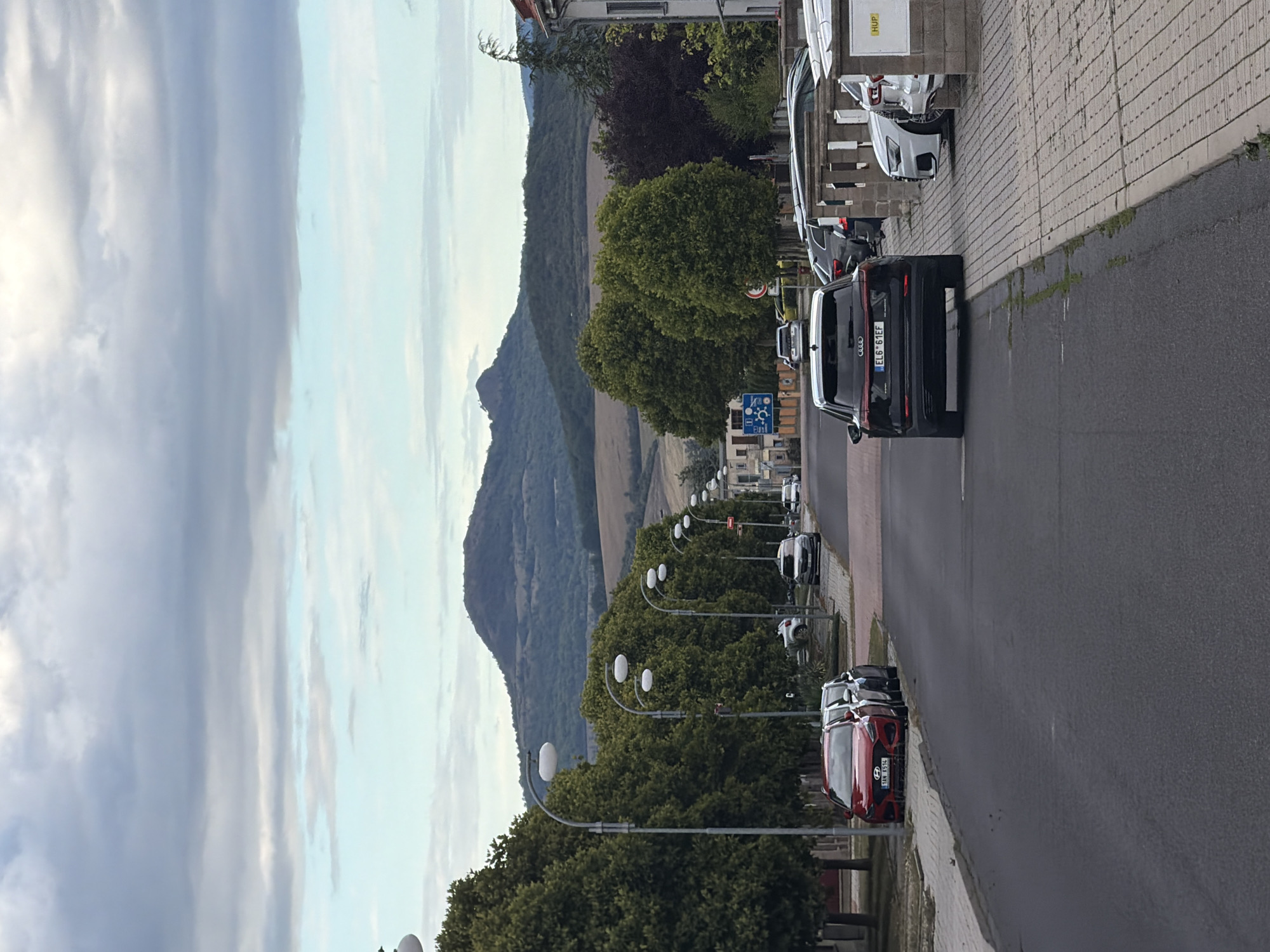 The image depicts a street scene in Louny, Czechia. A road runs through the center of the frame, lined with trees on the left and buildings and parked cars on the right. In the background, rolling hills and a prominent mountain range form the landscape under a cloudy sky. Several vehicles are visible, including a red Hyundai on the left and a black Audi in the foreground. Streetlights are spaced along the sidewalk. The overall impression is a quiet, overcast day in a European town.