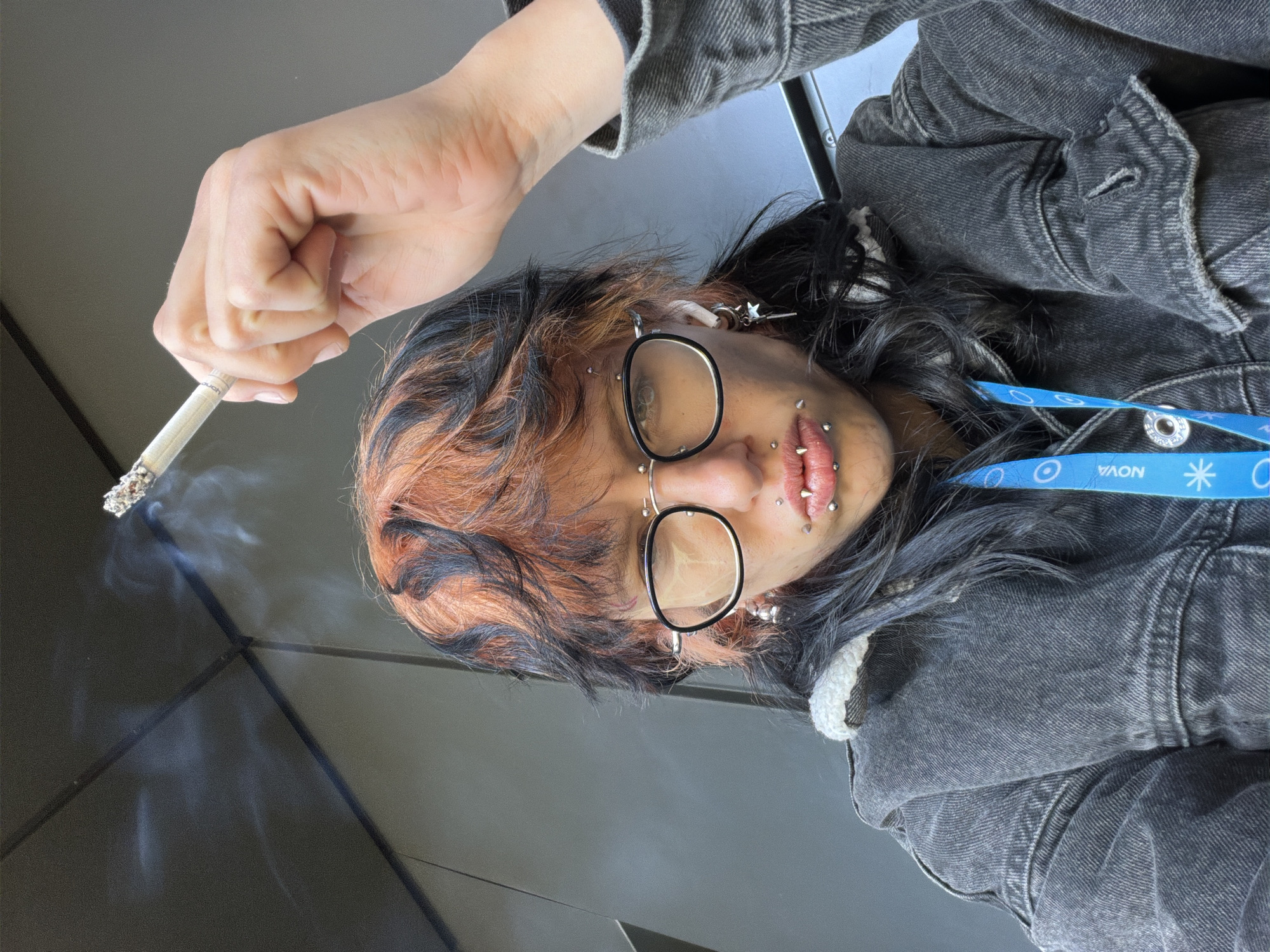 This close-up portrait, captured indoors in Busmantsi, Bulgaria, features a person holding a lit cigarette. The subject, viewed from a low, tilted angle, has dark hair with significant reddish-orange highlights. They wear round, black-rimmed glasses and multiple facial piercings, including studs and rings around the lips, nose, and eyebrow area. Small earrings, one featuring a star or key charm, are visible, as are in-ear headphones.
The person is dressed in a dark gray or black denim jacket, with a fluffy white lining visible at the collar or hood. Around their neck hangs a blue lanyard displaying the text "NOVA" repeatedly, alongside circular and asterisk-like logos. The lit cigarette, held by the person's hand, emits tendrils of smoke. Visible text on the cigarette filter includes "touch" and potentially "Rothmans."
The background is a simple, dark gray, likely an interior wall or floor, providing a stark contrast to the subject. The lighting is soft and even, suggesting an indoor setting without harsh direct light. The subject's gaze is direct, and their expression is neutral.