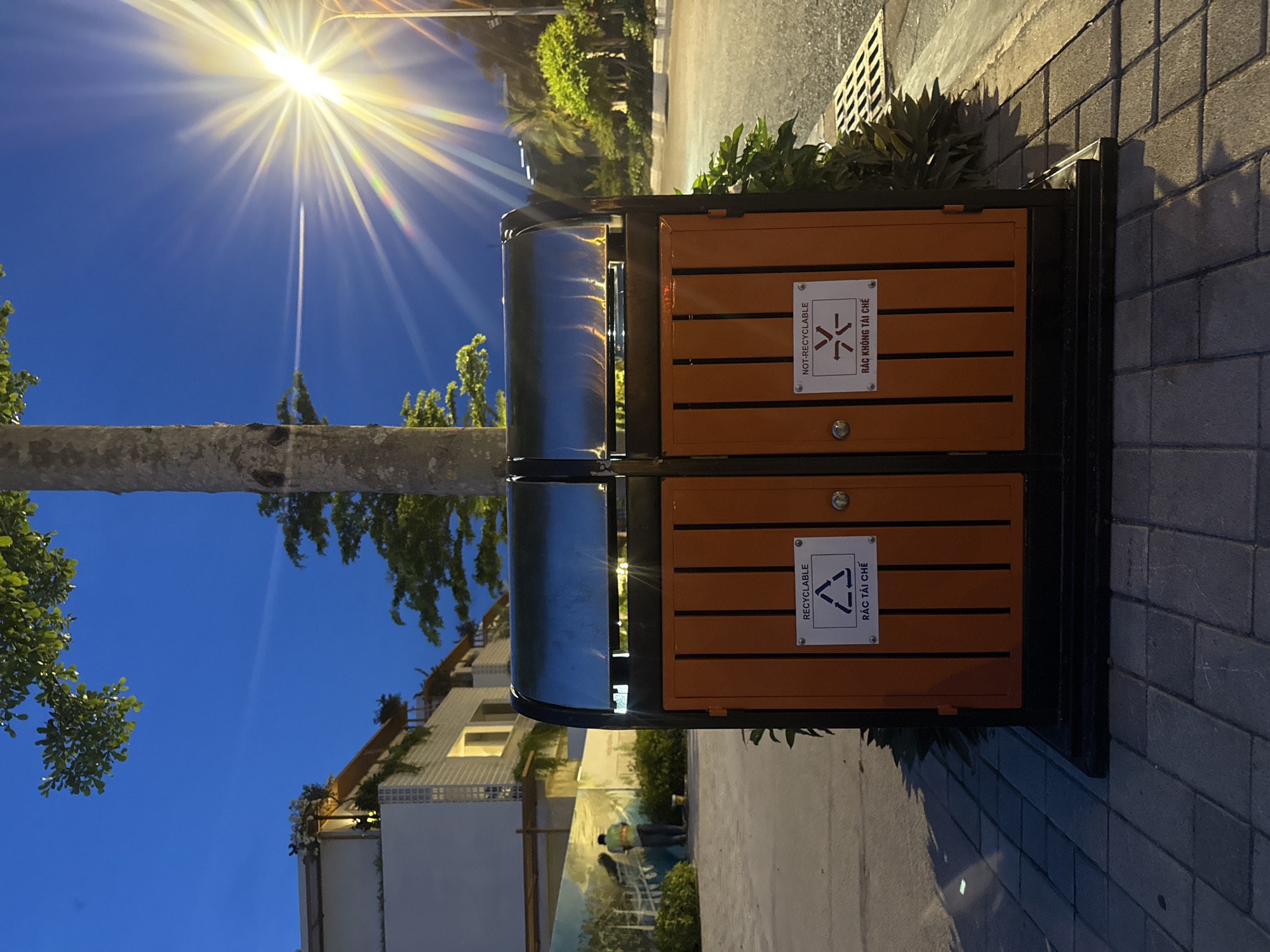 In the city of Bac Nha Trang Ward, Vietnam, a dual-compartment outdoor trash receptacle stands on a paved walkway at dusk. The receptacle features two orange doors, each with a white sign. The left door is labeled "RECYCLABLE" with a recycling symbol and Vietnamese text. The right door is labeled "NOT-RECYCLABLE" with a red "X" over the recycling symbol and Vietnamese text. The receptacle is positioned in front of lush green foliage and a tree trunk. In the background, a white building with multiple levels and illuminated windows is visible, along with a streetlamp casting a bright glow. A person in a yellow vest can be seen in the distance, possibly painting a mural on a wall. The sky is a deep blue, indicating twilight.