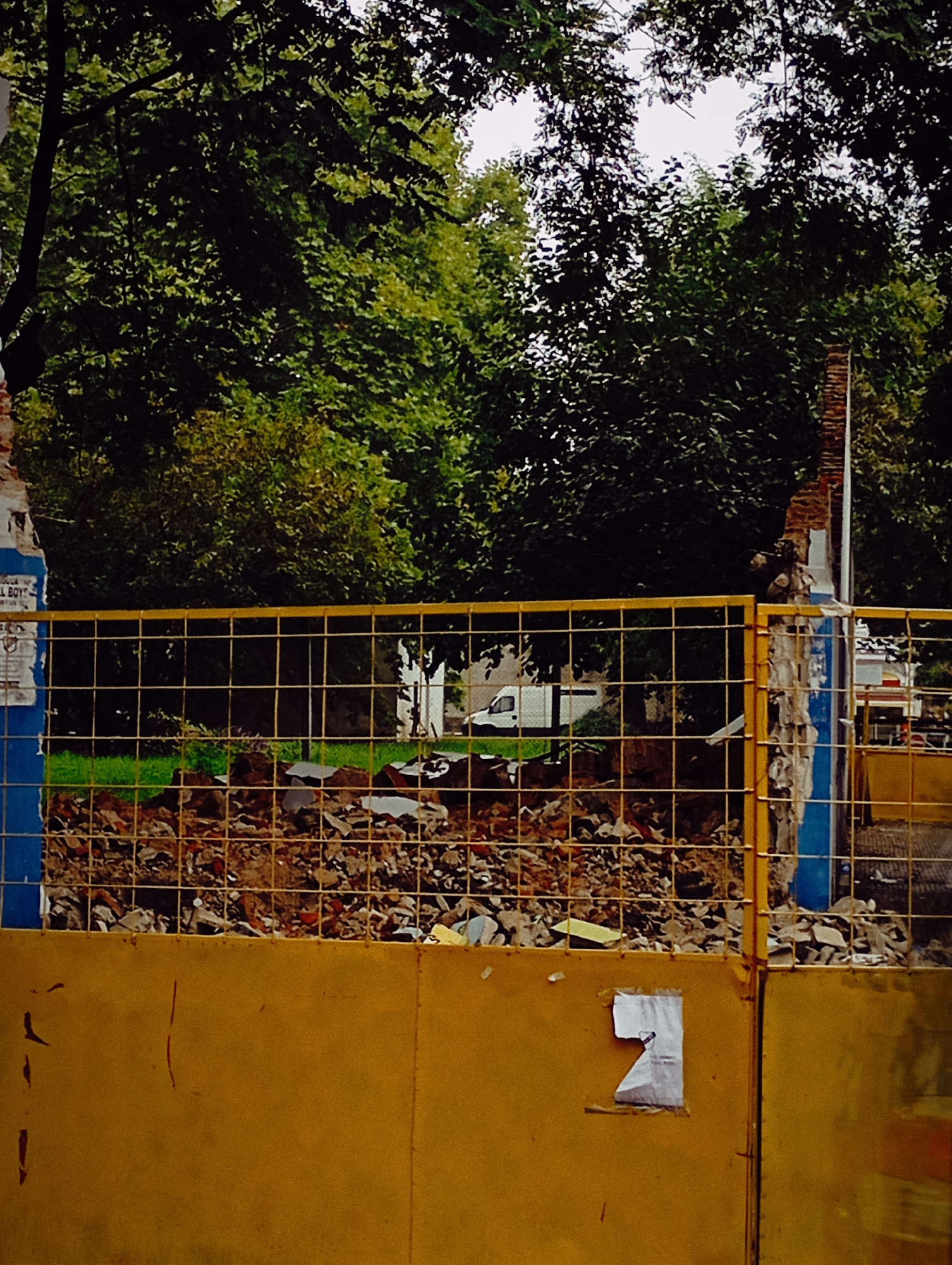 The image shows a construction or demolition site in Buenos Aires, Argentina, viewed through a yellow metal fence with a wire mesh upper section. Debris consisting of broken brick and concrete is visible behind the fence. The trees are in the background, partially obscuring some vehicles in the far distance. One piece of paper is loosely taped to the yellow fence. The fence is painted with blue vertical elements and a yellow horizontal element on the corners.