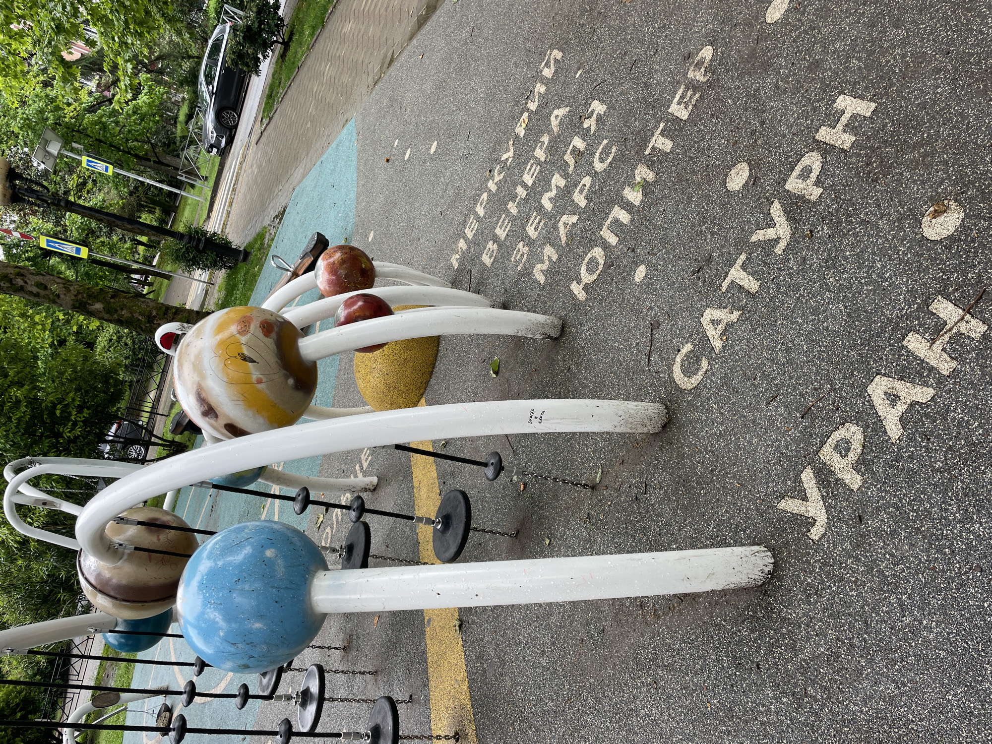 The image depicts a model of the solar system in a playground, likely located in Sochi, Russia based on the Cyrillic lettering of the planet names (Меркурий, Венера, Земля, Марс, Юпитер, Сатурн, Уран). The model features painted spheres representing the planets mounted on a metal structure. The planet models are connected by chains and discs. The background includes trees, a road, and a car, suggesting an urban park environment. The ground is paved with the planet names painted in white.