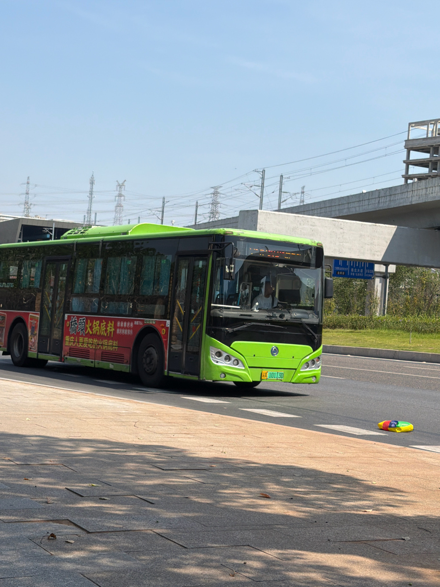A bright green city bus is traveling on a road in Jiangjin, China. The bus has advertisements on its side. A driver is visible inside the bus. The bus's license plate reads "A 101 3D". In the foreground, a sidewalk is paved with gray stones. A colorful toy is lying on the ground near the sidewalk. In the background, there are power lines and elevated structures, indicating ongoing construction or infrastructure. The sky is clear and blue.