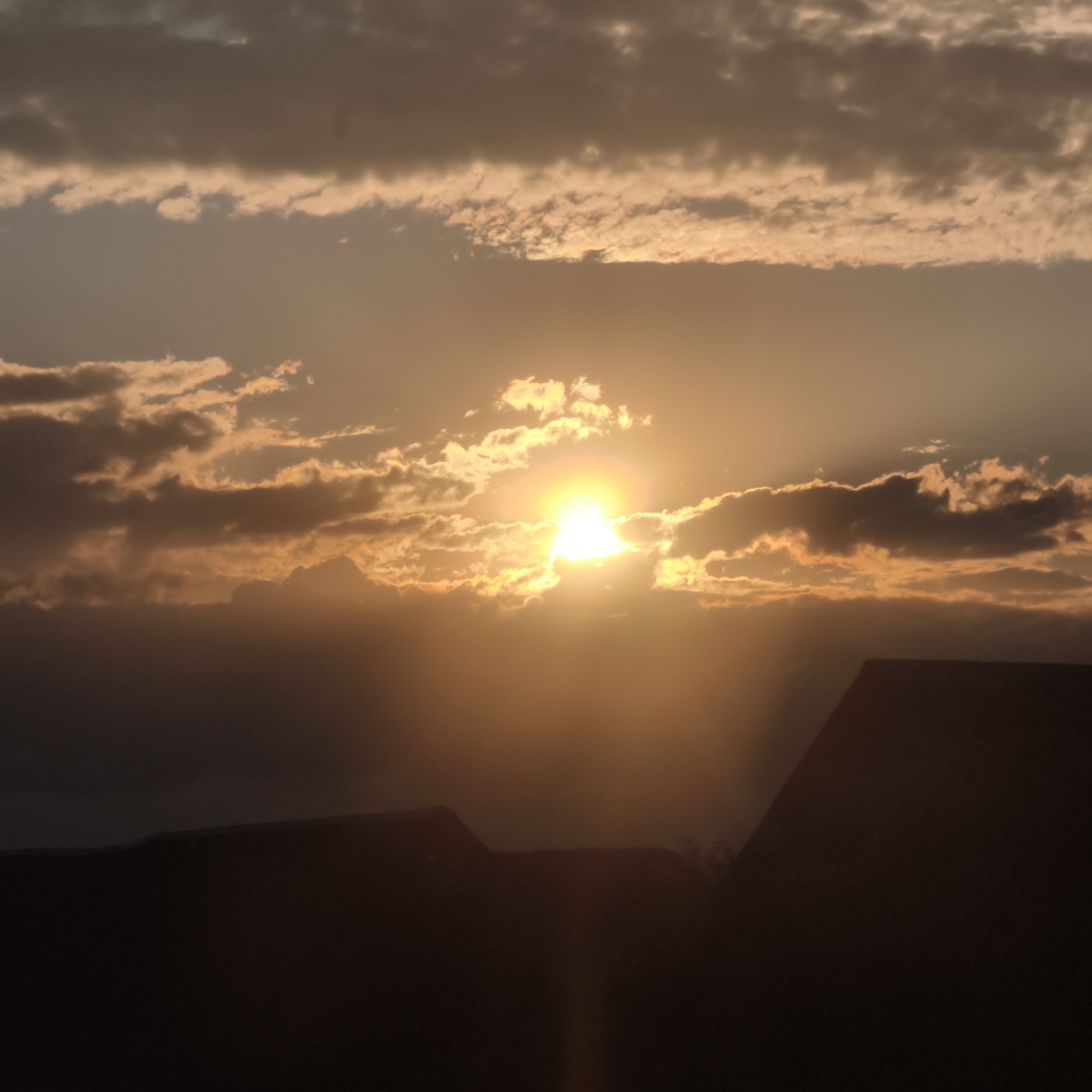 The image depicts a vibrant sunset or sunrise scene dominated by warm hues and dramatic cloud formations. A bright, intensely glowing sun is positioned centrally, partially obscured by dark, broken clouds. Rays of light, known as crepuscular rays, emanate from behind the clouds, casting a radiant glow across the sky. The clouds themselves are varied, with darker, thicker layers interspersed with lighter, orange-illuminated sections reflecting the sun's light. The upper portion of the sky shows subtle shifts in color from a soft, hazy orange near the horizon to a muted gray-blue higher up. In the foreground, the dark silhouettes of what appear to be multiple rooftops or building structures are visible, suggesting an urban or suburban environment. The scene evokes a sense of natural beauty and tranquility. There are no people or discernible text in the image.