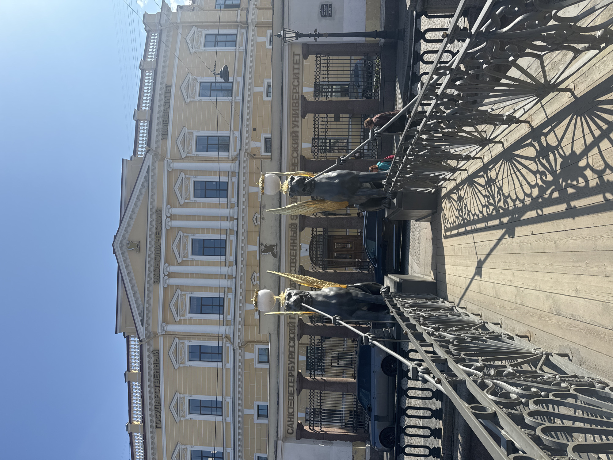 This media file captures a sunny daytime scene on a bridge in Saint Petersburg, Russia. The image is oriented vertically (rotated 90 degrees clockwise).
In the foreground, a decorative metal bridge railing stretches across the frame, casting intricate shadows onto a wooden surface, likely the bridge deck. Two iconic griffins, mythical creatures with dark bodies, golden wings, and holding spheres with lamps, are prominently featured on the railing. Their ornate design is a notable detail.
Behind the bridge and its statues stands a grand, pale yellow neoclassical building with multiple windows, white columns, and decorative cornices. Visible text on the building facade clearly identifies it as "САНКТ-ПЕТЕРБУРГСКИЙ ГОСУДАРСТВЕННЫЙ ЭКОНОМИЧЕСКИЙ УНИВЕРСИТЕТ" (Saint Petersburg State University of Economics). Another sign on the building indicates "ГРИБОЕДОВА 30-32," which is the address (Griboyedov Canal Embankment).
Two individuals are partially visible on the bridge behind the griffins, suggesting pedestrian activity. A traditional lamppost stands near the building entrance. Several cars are parked or passing by the building's base. The sky is bright blue and clear, indicating excellent weather conditions and strong sunlight, which creates crisp shadows. The overall setting evokes a sense of historical architecture and bustling urban life.