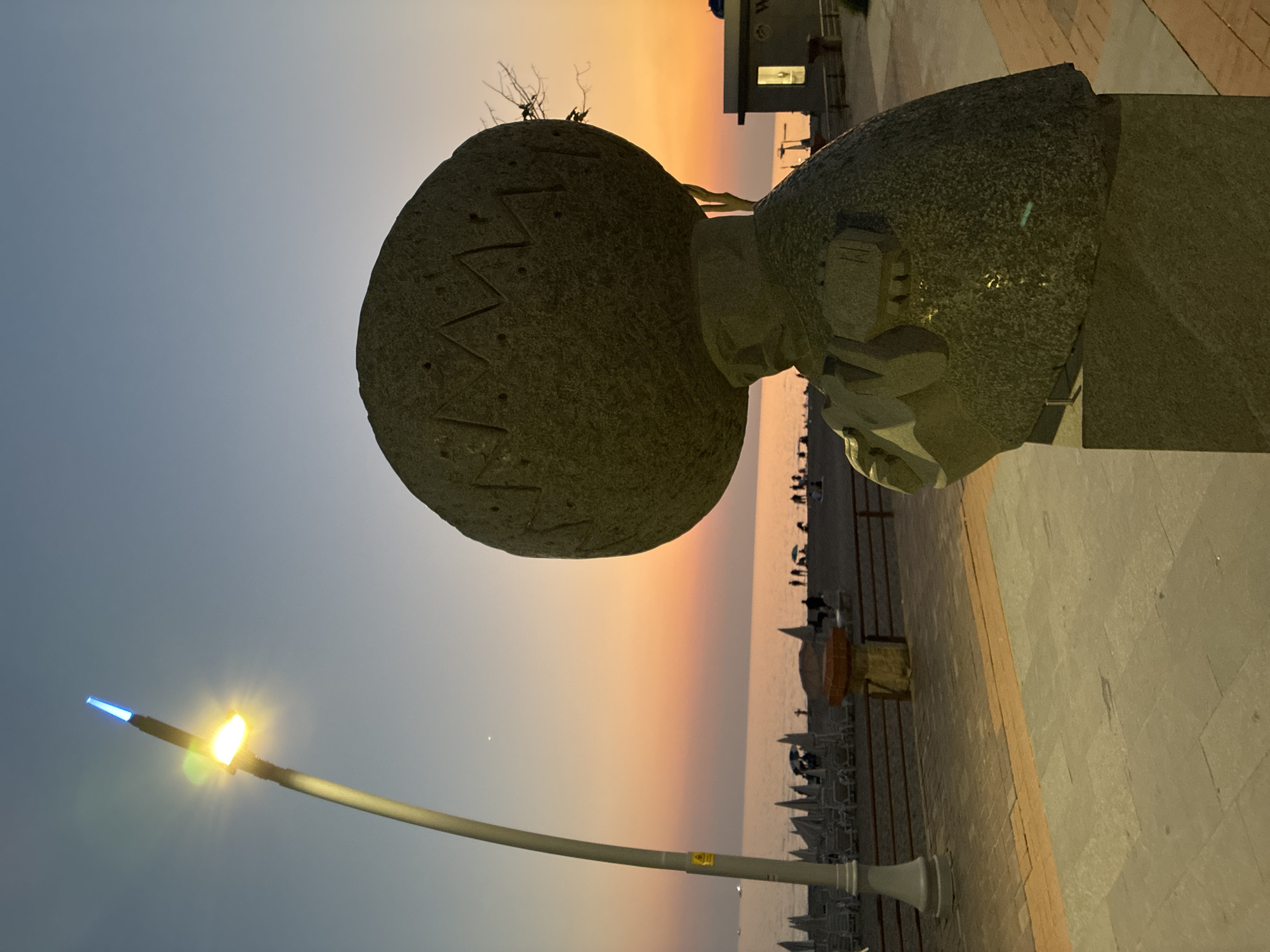 A statue of a figure with a large, spherical head stands on a paved promenade by the sea. The statue is made of dark, rough-textured stone and is positioned in the foreground on the right side of the image. The figure's head is adorned with carved patterns. Behind the statue, a beach with rows of lounge chairs and umbrellas is visible, leading to the calm sea. The sky displays a gradient of colors, transitioning from a pale blue at the top to soft orange and pink hues near the horizon, indicating sunset or sunrise. A street lamp with a bright yellow light and a blue-tipped beacon stands on the left side of the promenade. Silhouettes of people are scattered across the beach and in the water. A small building with a lit window is present on the right side of the scene, further down the promenade. The overall environment suggests a coastal resort area during twilight.