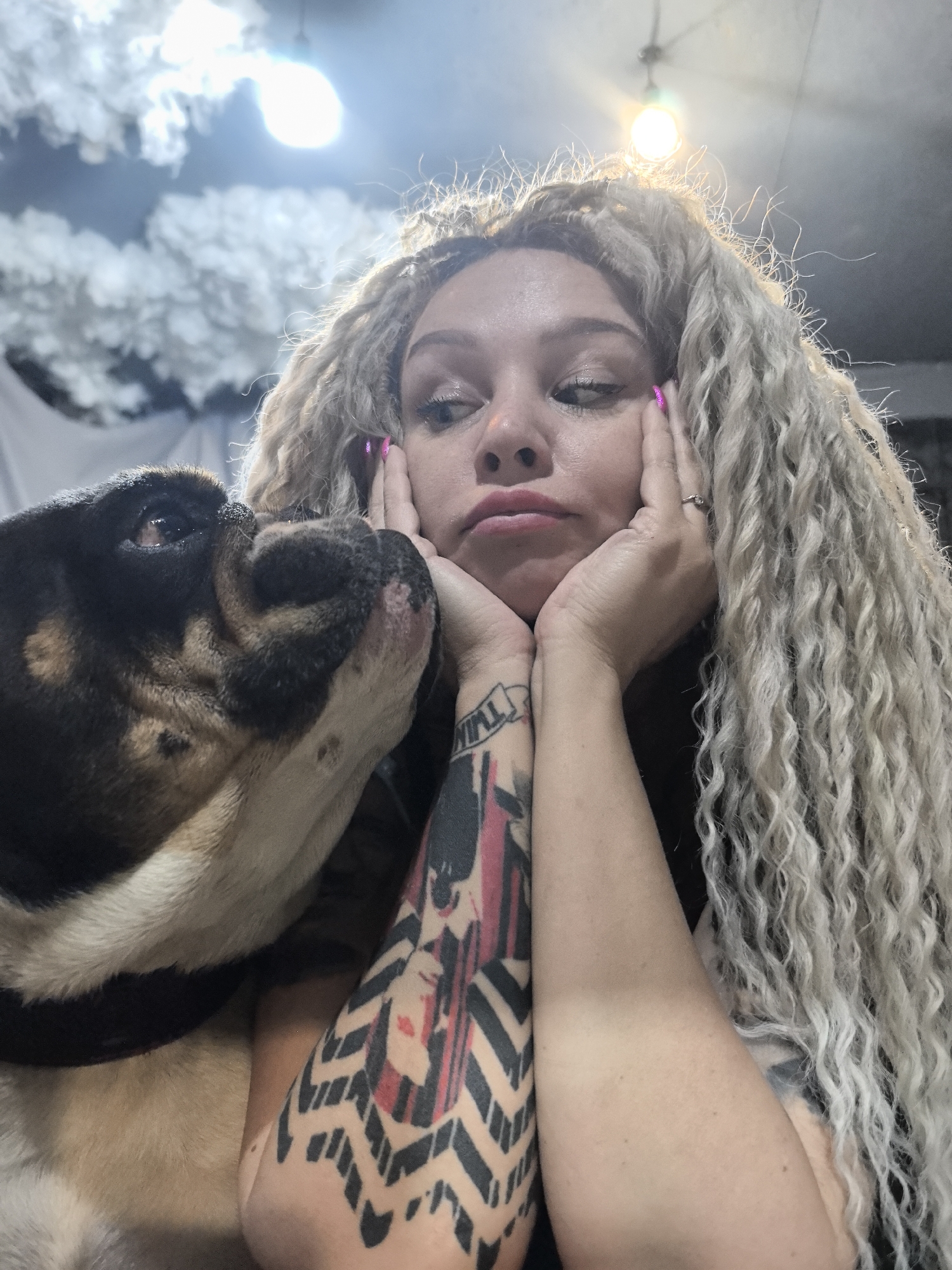 A woman with long, wavy blonde hair and a tattoo on her left forearm is holding her face with her hands. She has pink fingernails and appears to be pouting. A black and tan bulldog is nuzzling her face. The background includes artificial clouds and lighting fixtures. The location is not discernible from the image provided.