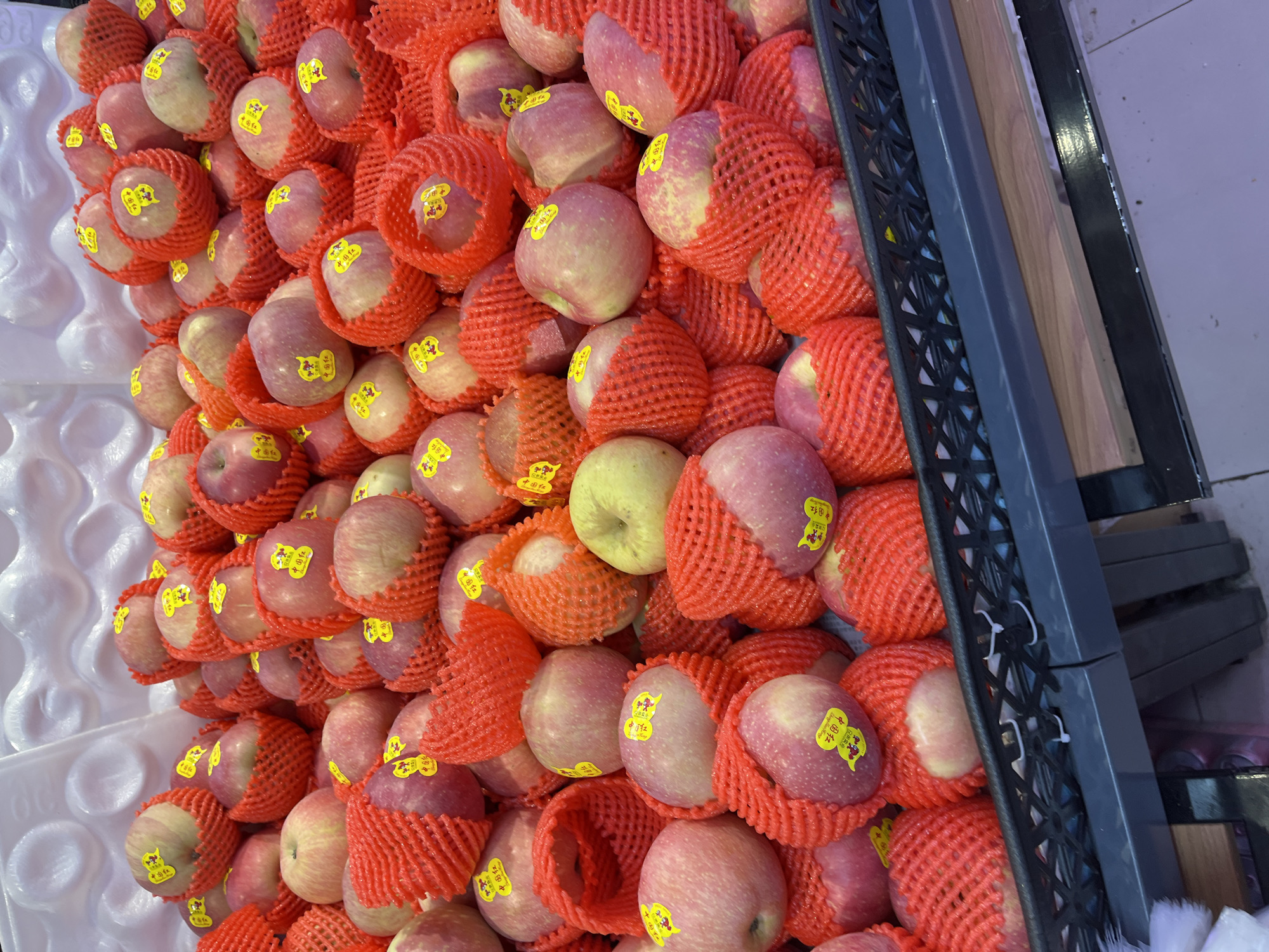 Here's a detailed description of the image:
The image shows a large pile of red apples, each individually wrapped in a red protective mesh. Many of the apples have a yellow sticker on them featuring Chinese characters; the text "中国红" (Zhōngguó Hóng) is visible on the stickers, translating to "China Red." The apples are packed closely together in a black plastic tray or container, with part of the tray visible on the right side of the image. The left side of the image shows a white styrofoam-like material, potentially used for cushioning or display. The setting appears to be a store or market, presumably in Changchun City, China.