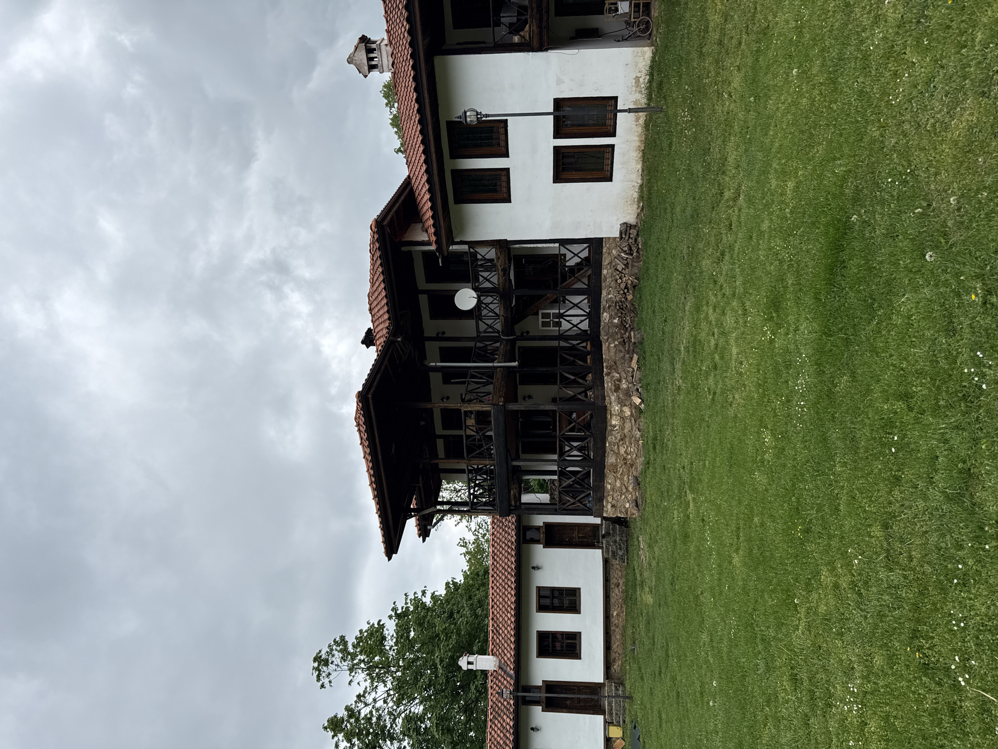 The image shows a two-story building with a red tiled roof and white walls, located in Etropole, Bulgaria. The building features wooden balconies with dark brown supports and railings. There are windows with dark frames on both floors. The facade is white with a stone foundation. A satellite dish is mounted on the balcony. A chimney rises from the roof. The building is surrounded by a green lawn. The sky is overcast with grey clouds. A tree is visible near the left side of the building. A lamppost is located in front of the building. There are logs of wood stacked next to the wall.