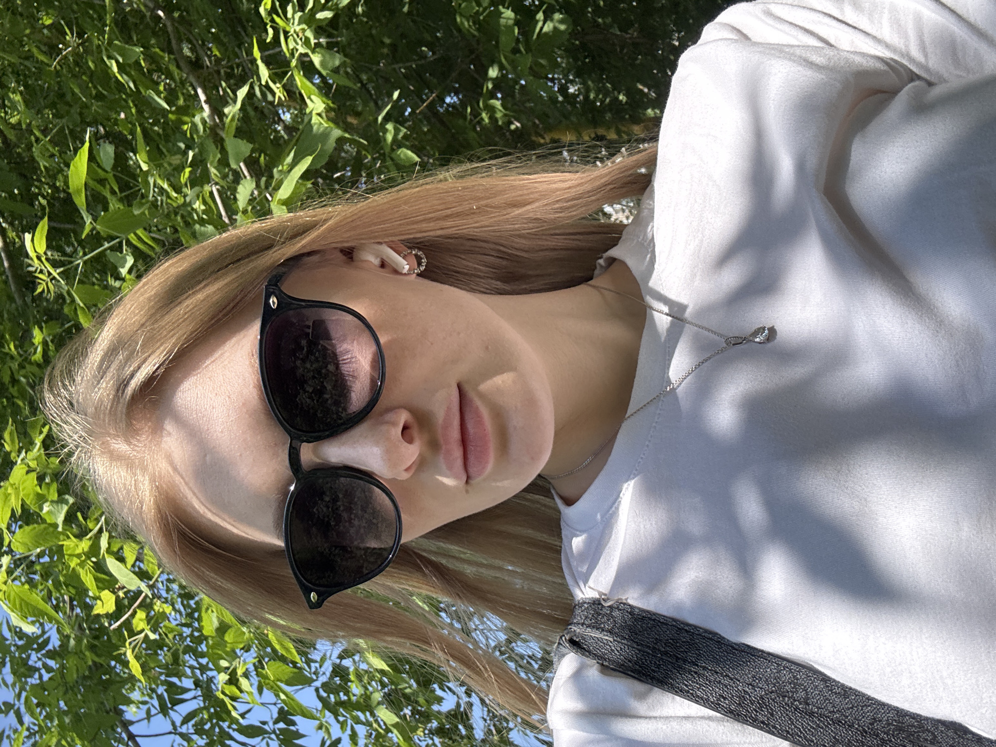 A young, light-skinned woman with blonde hair and sunglasses is photographed outdoors in daylight. She is wearing a white top with a dark-colored shoulder strap or bag strap visible. A silver necklace with a pendant is around her neck. Behind her, foliage and blue sky can be seen.
No details present allow to confirm the location in the city of Perm, Russia.