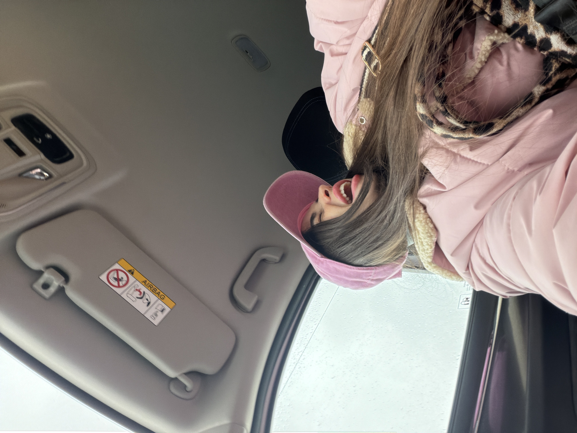 A woman wearing a pink baseball cap and a pink puffer jacket with a leopard print collar is inside a car. The interior of the car includes a gray sun visor with an airbag warning sticker, a gray ceiling with a console, and a black seat. Raindrops are visible on the car window. The location is unspecified; there is no evidence to indicate the city of Saint Petersburg, Russia.