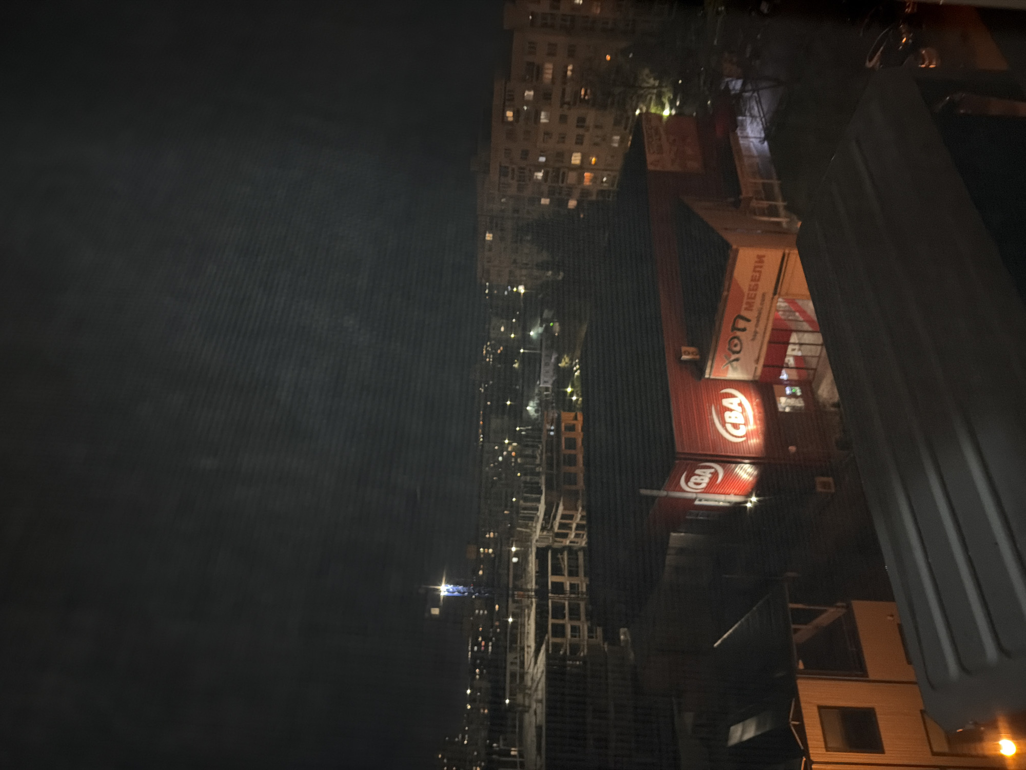 The image shows a nighttime urban view, likely from a high vantage point in Sofia, Bulgaria. In the middle, multiple buildings of varying heights and states of construction stand. Building facades are lit up with a mix of windows. The bottom right of the image includes a section of a white truck. A red store with the letters "CBA" is visible near the center. The left side of the image is dark, obscuring details.