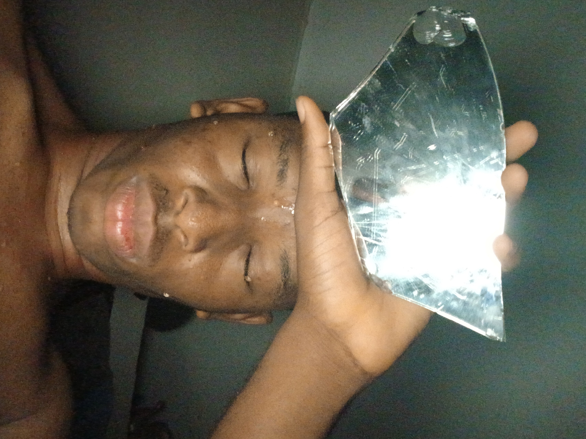 The image shows a person with dark skin, likely male, positioned on their side with their eyes closed. Drops of liquid, appearing to be water or sweat, are visible on their forehead and around their mouth, which is slightly parted. The person's right hand is raised, holding a jagged piece of broken mirror near their head. The mirror's surface is scratched and reflects a prominent bright light source, casting a strong glare. The background is dark and indistinct, suggesting a dimly lit indoor environment. No text is visible. The scene conveys a somber or contemplative mood. This image is set in Lagos, Nigeria.