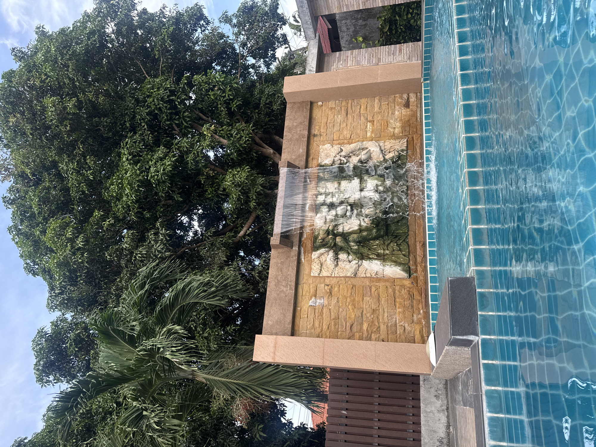 A swimming pool in Baan Bo Phut, Thailand, features a waterfall cascading over a stone-tiled wall with bas-relief carvings. The pool's blue tiles reflect the clear sky and surrounding greenery. A wooden fence lines the left side of the frame, while lush trees, including a palm tree, dominate the background. The scene is tranquil and inviting, indicative of a resort or private residence in a tropical setting. There are no people or animals visible in the image.